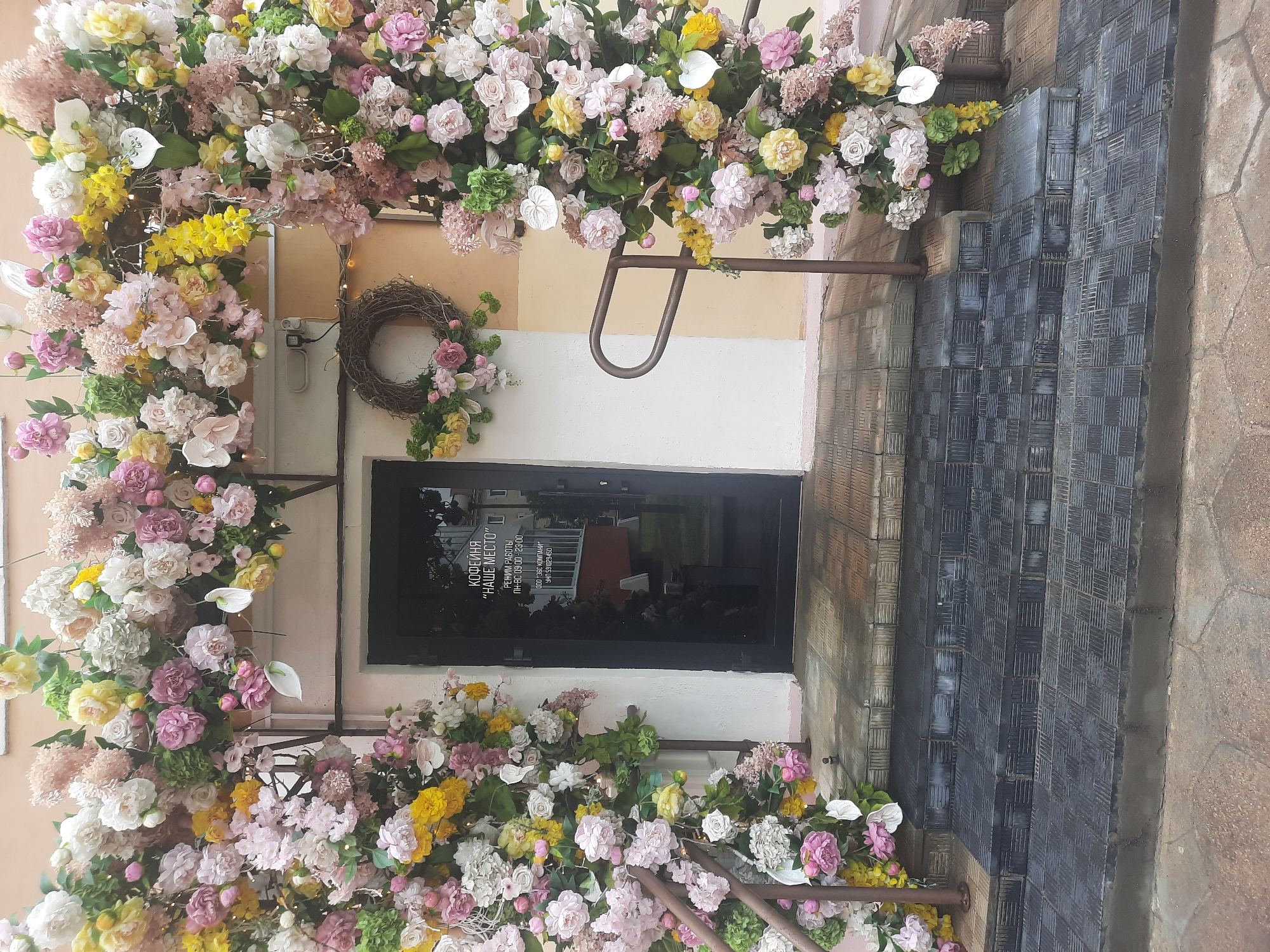 The image depicts the entrance to a business, likely a cafe or coffee shop, adorned with an elaborate floral archway. The archway is constructed from a dense arrangement of artificial flowers in various pastel shades of pink, yellow, and white, with green foliage interspersed throughout. Tiny fairy lights are woven into the floral display, particularly on the right side.
On the left side of the entrance, a rustic wreath made of twigs and decorated with a few small pink flowers hangs on the wall. To the right of the wreath and beneath the floral arch, a metal railing runs alongside the stairs. The entrance itself is a dark glass door with text superimposed on it. The text, in Russian, reads "КОФЕЙНЯ \"НАШЕ МЕСТО\"" which translates to "COFFEE SHOP \"OUR PLACE\"". Below this, it indicates operating hours: "РЕЖИМ РАБОТЫ ПН-ВС 09:00 - 23:00" (Working hours Mon-Sun 09:00 - 23:00). Further down is company information: "ООО \"ЭБС КОМПАНИ\"" and "УНП 591029450".
The entrance is accessed via a set of stairs, with a ramp to the right of the stairs. The stairs and ramp are covered with dark, textured rubber tiling. The building facade appears to be painted in muted tones of cream and a light peachy-yellow. The ground in front of the entrance is paved with stone tiles. The overall impression is one of a festive and inviting storefront decoration. This scene is located in the city of Hrodna, Belarus, as indicated by the Russian text on the door and the general architectural style. There are no people visible in the image.