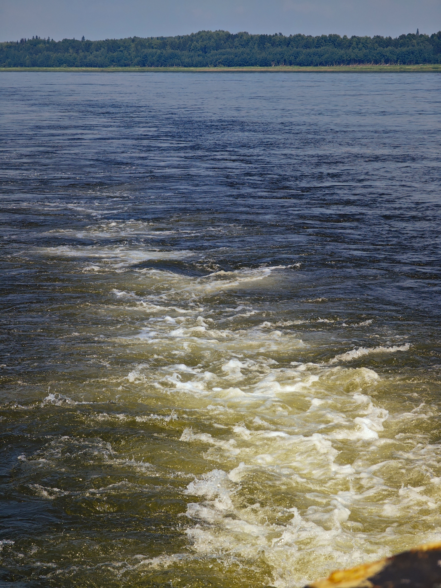 A wide shot captures a turbulent expanse of dark blue water, leading to a frothy, golden-brown wake in the foreground. The wake, characterized by swirling foam and disturbed water, suggests movement, possibly from a vessel. In the distance, a dense, green treeline stretches across the horizon under a pale, overcast sky. The scene appears to be a natural body of water, such as a river or lake, with no visible structures or individuals. The location is the Pakhra River in the village of Pogodaevo, Russia.