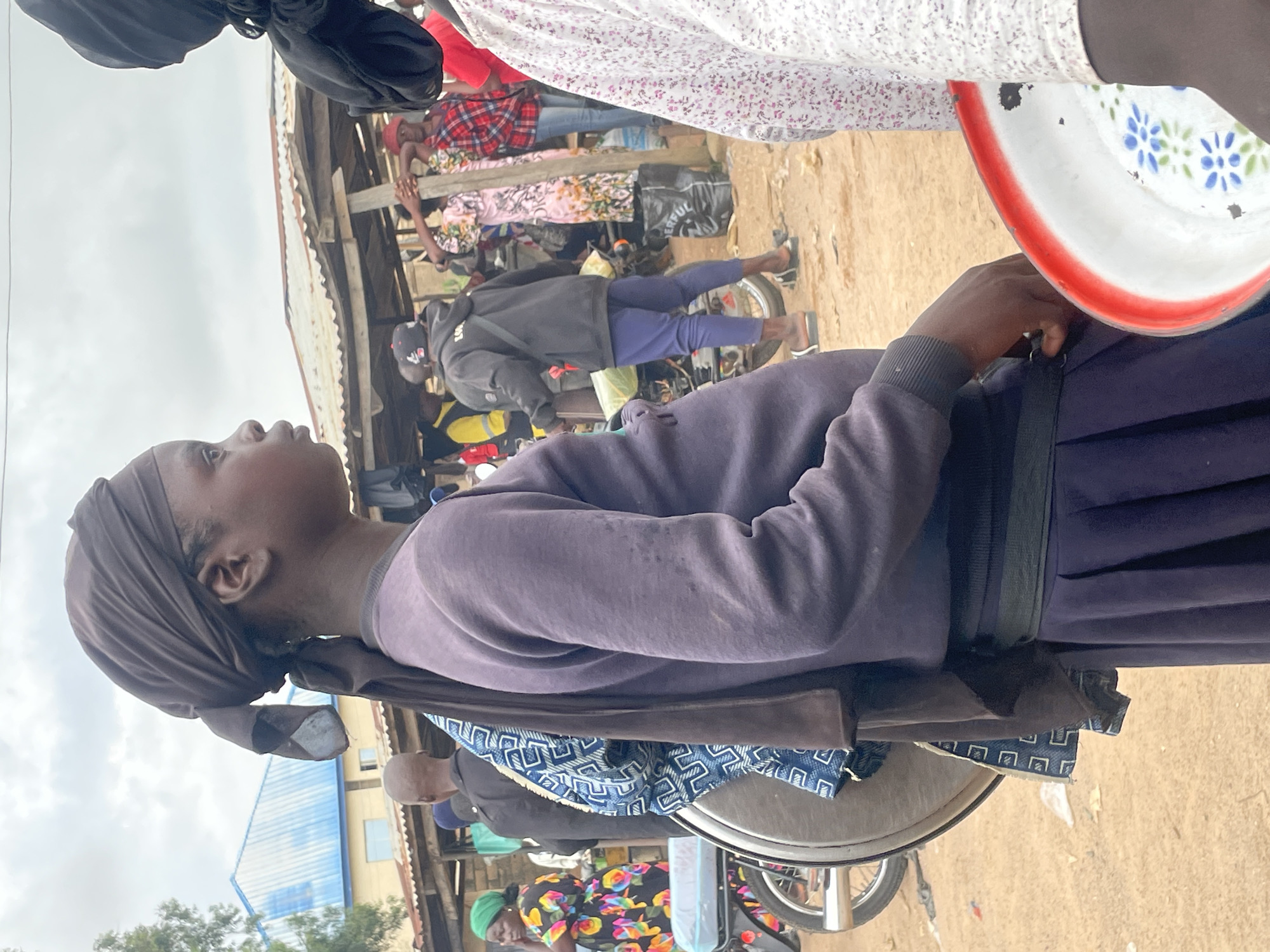 A young Black female wearing a dark brown headscarf and a long-sleeved purple sweater is standing in what appears to be a market in Wang, Nigeria. She is looking to her right, with her mouth slightly open. Her hair is braided and tucked under the headscarf, with a portion of the scarf draped over her shoulder. She is wearing a dark purple pleated skirt. To her left, a silver circular object, possibly part of a drum or large container, is visible. A blue and white patterned fabric is draped over this object. In the background, there are several other people, some standing under wooden stalls with corrugated metal roofs, suggesting a bustling market environment. The ground is dusty and appears to be made of dirt. The sky is overcast. Other individuals in the background are wearing various clothing, including a woman in a brightly colored floral dress and a man in a black hoodie. To the right of the main subject, a white basin with a red rim and a blue floral pattern is being held by an unseen person.