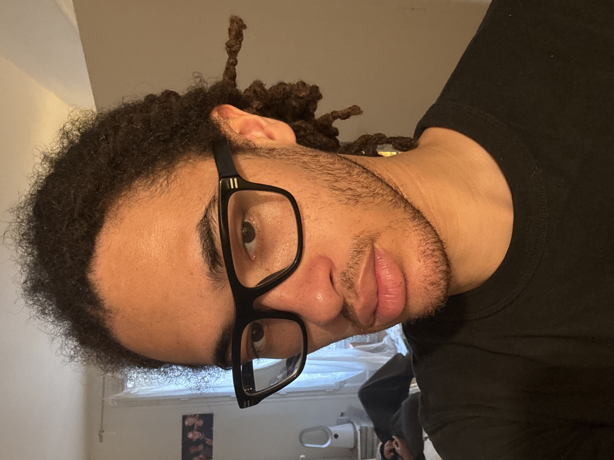 A close-up, slightly rotated image features a man with dark curly hair and dreadlocks, wearing black-framed glasses and a black t-shirt. He has light skin and a neatly trimmed beard and mustache. The background shows a glimpse of a room with white walls, a window, and what appears to be a toilet. There is also a person in the lower background, but the details are unclear. There is no evidence to indicate that this photo was taken in Paris, France.