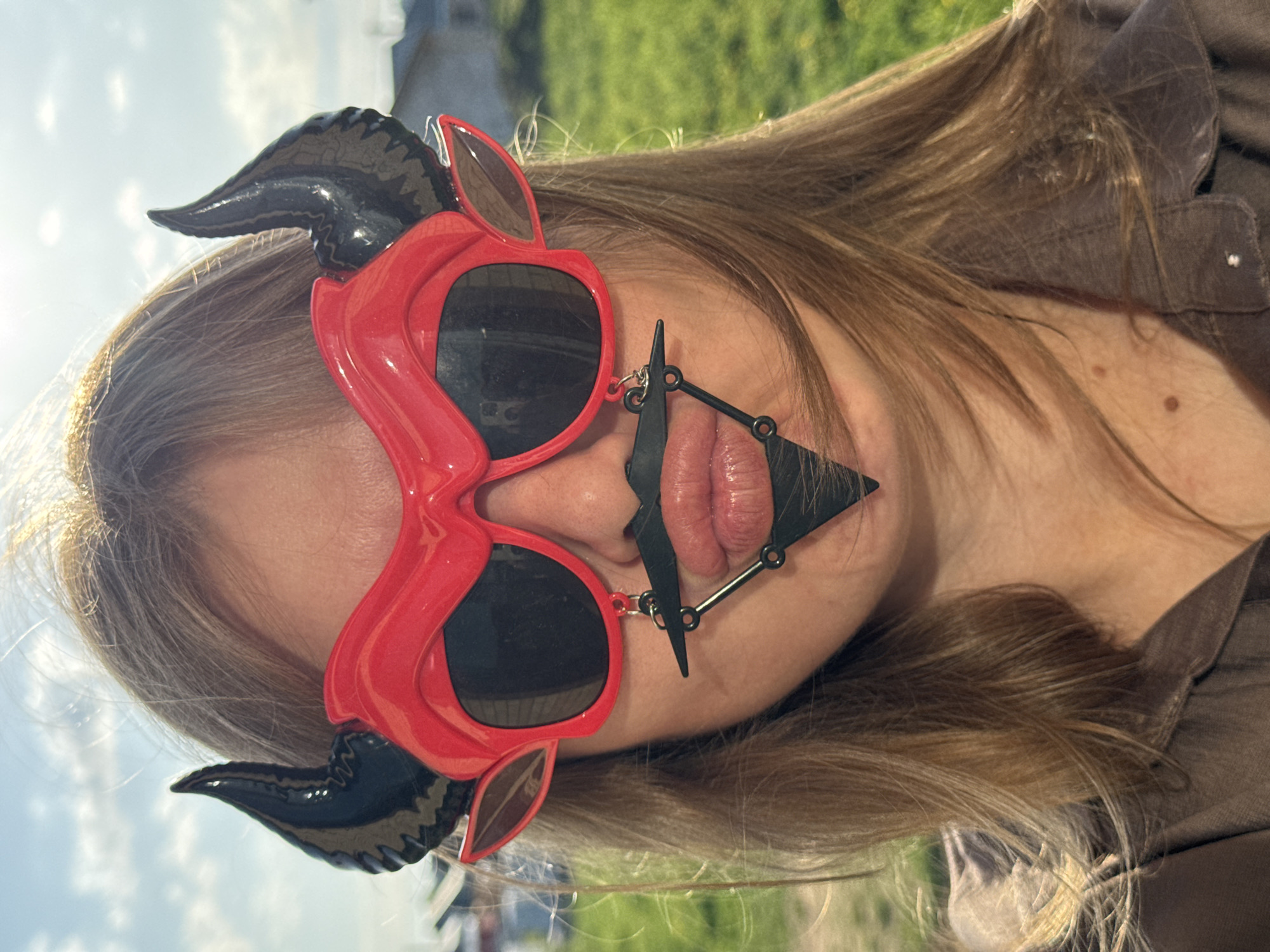The image shows a woman with long, light brown hair, wearing red sunglasses with black horns attached to the sides. She is also wearing a black triangular object attached to her lip with chains and rings. The background is blurred but appears to be an outdoor setting with green grass and trees, possibly taken in the city of Ферма, Russia. She is wearing a brown shirt. The photo appears to be taken at an angle.