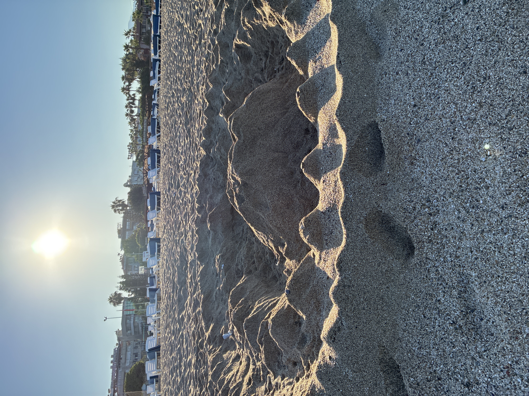 The image depicts a sandy beach in Alanya, Turkey, with the sun shining brightly in a clear blue sky. In the foreground, a circular structure of sand has been built, with smaller mounds of sand forming a textured pattern within and around it. In the middle ground, rows of empty white lounge chairs with blue cushions are arranged on the sand, some covered by light blue awnings. Beyond the chairs, a line of buildings and palm trees can be seen, with some taller structures indicating hotels or residential buildings. The overall scene suggests a peaceful, early morning or late afternoon on a well-maintained beach resort. The light suggests the sun is low in the sky, casting long shadows across the sand.