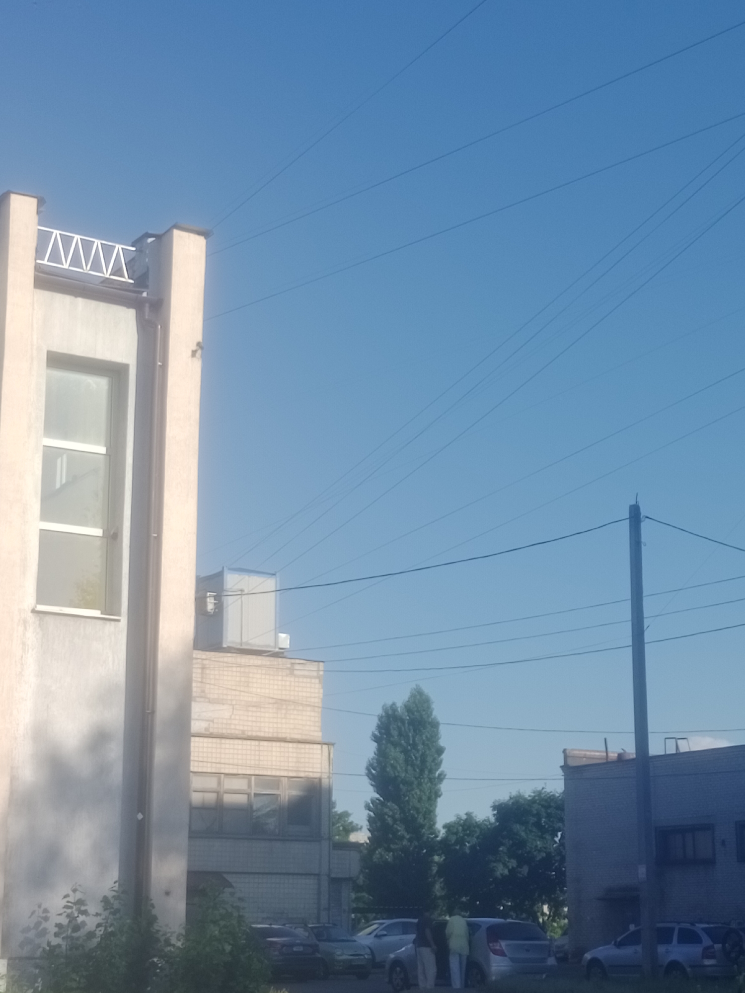 The image depicts an outdoor scene in Horishni Plavni, Ukraine, on a clear, bright day under a blue sky. In the foreground on the left is a light-colored building with a prominent vertical pillar and a large window with multiple panes. The building appears to be two stories high. To the right of this building is another, taller structure made of light-colored bricks, with multiple windows visible. The top of this brick building features a white, box-like structure. In the distance, a tall, slender evergreen tree stands to the right of the brick building, with other trees and foliage surrounding it. Several parked cars are visible in the middle ground, including a dark-colored sedan and a silver hatchback. Three individuals are standing near the silver hatchback, with one person wearing a yellow garment. Further to the right, another brick building is visible, appearing to be a single-story structure with a few windows. A wooden utility pole with electrical wires extending across the sky is prominent in the right side of the frame. The presence of overhead wires crisscrossing the sky suggests an urban or industrial setting.