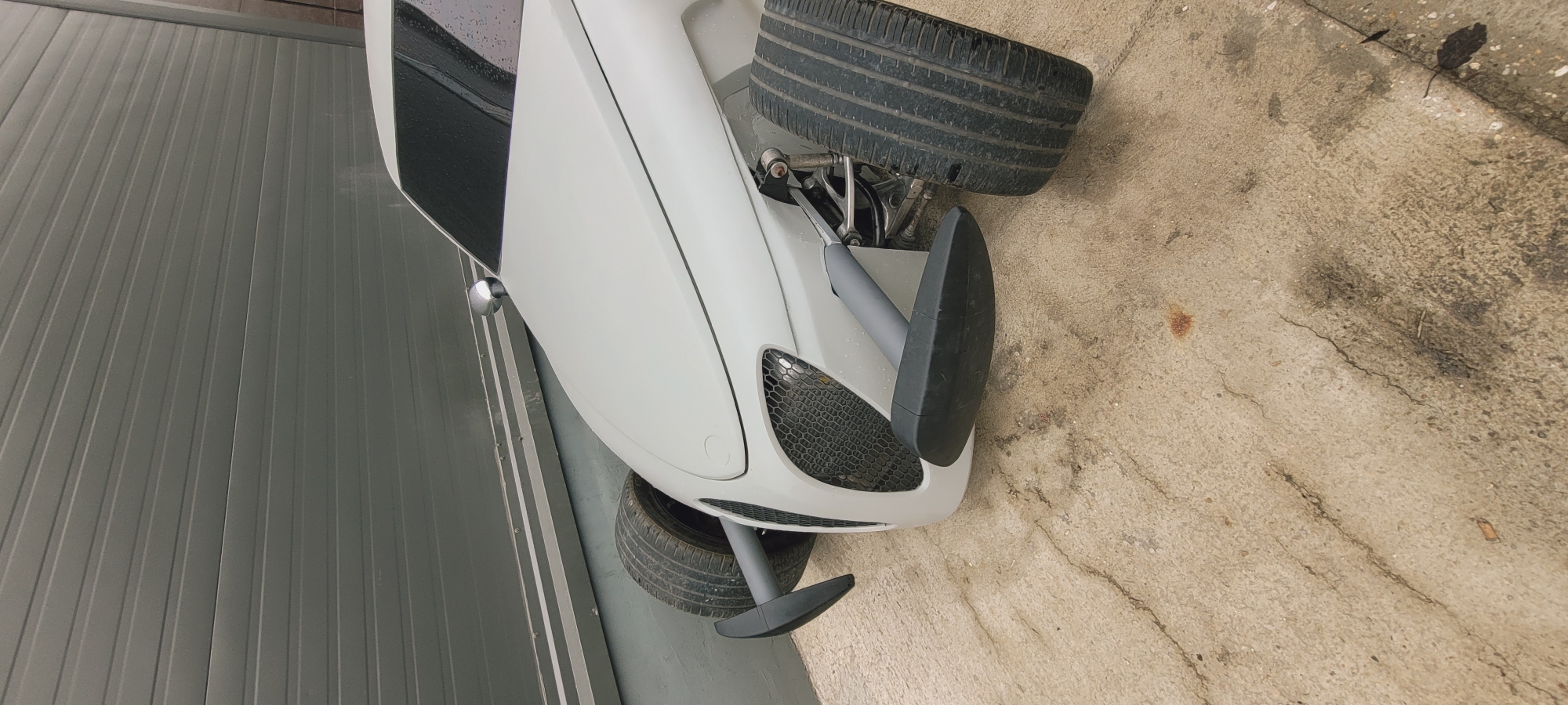 A car is parked beside a metal wall on a concrete surface in an urban location in Unknown, Unknown. The front of the vehicle is positioned towards the viewer, revealing two tires and suspension components. The car's body is white with a black air vent on the left side. A chrome mirror is visible on the left side of the car's body. The concrete surface is light brown with cracks and stains. The metal wall is gray and vertical.