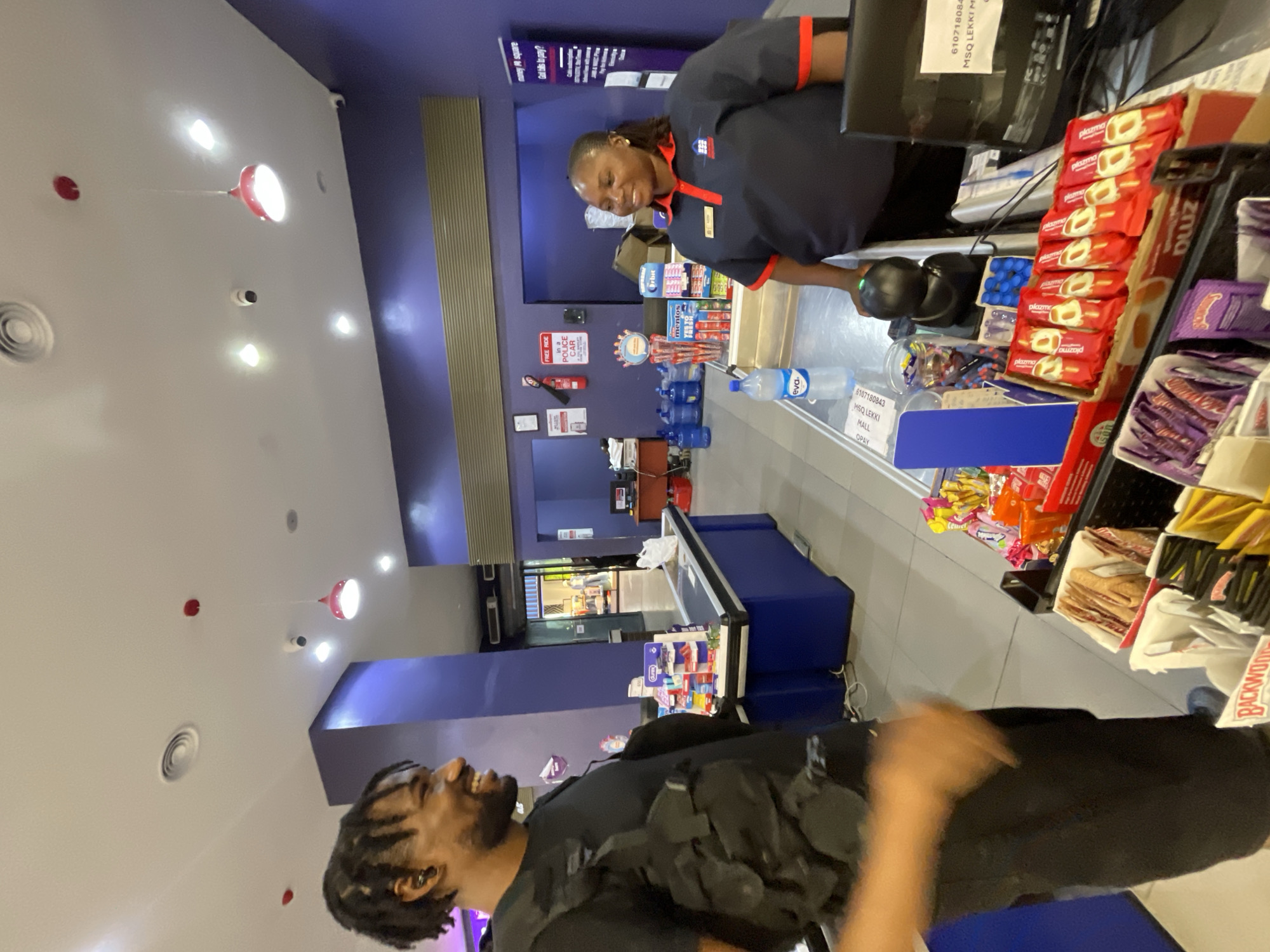 The image depicts an indoor scene at a retail establishment, likely a convenience store or supermarket, located in Moba, Nigeria. A Black male customer, wearing a black t-shirt and a tactical vest, stands on the left side of the frame, facing a female cashier. He has dreadlocks and is smiling. The cashier, who appears to be of average build with dark skin and hair pulled back, is wearing a dark blue polo shirt with red accents and a nametag. She is standing behind a blue counter, looking towards the customer and smiling.
The counter is filled with various products, including rows of wrapped ice cream bars, candies, and bottled water. A scanner and a computer monitor are visible on the counter. In the background, shelves stocked with goods can be seen, and a wide entrance with open doors leads to another section of the store. The walls are painted in a vibrant purple. The ceiling features recessed lighting and some ventilation fixtures. The floor is tiled. The overall environment suggests a commercial transaction taking place.