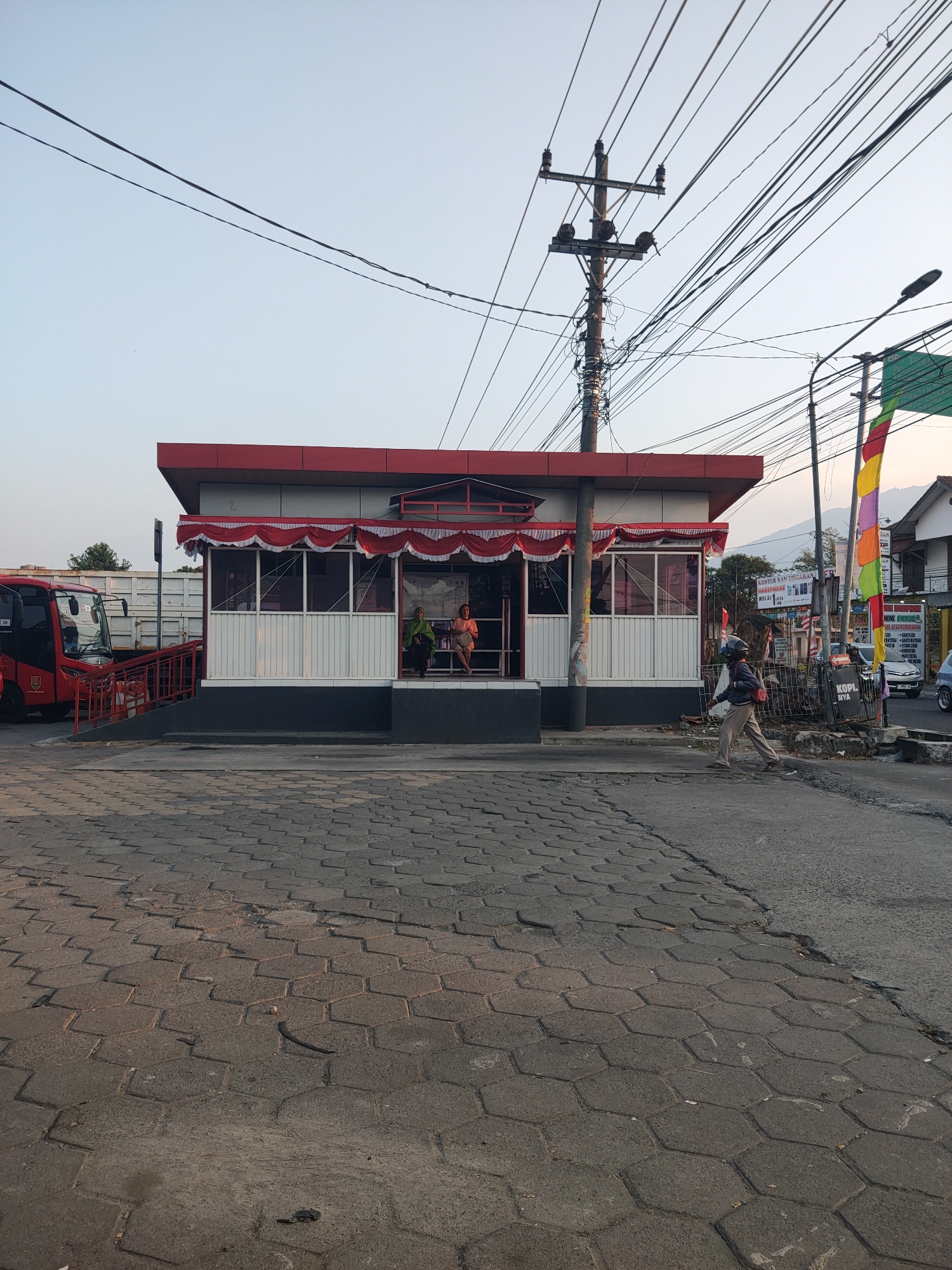 The image captures a scene in Ungaran, Indonesia, likely near a transportation hub. A small, modern-looking red and white building with a covered porch area, adorned with red and white festive fabric, stands prominently. Two women are seated on a bench inside the covered area. To the left, a red bus is parked, and a ramp leads up to the building's entrance. A person wearing a helmet and backpack is walking across the paved road in front of the building. Overhead, a dense network of electrical wires crisscrosses the sky, supported by a utility pole. Colorful flags, including a yellow, red, and green one, are visible on the right side of the frame, along with signage for businesses, including "Kopi. Nya." The ground is paved with hexagonal interlocking concrete blocks. The sky is clear and pale, suggesting it's either early morning or late afternoon.