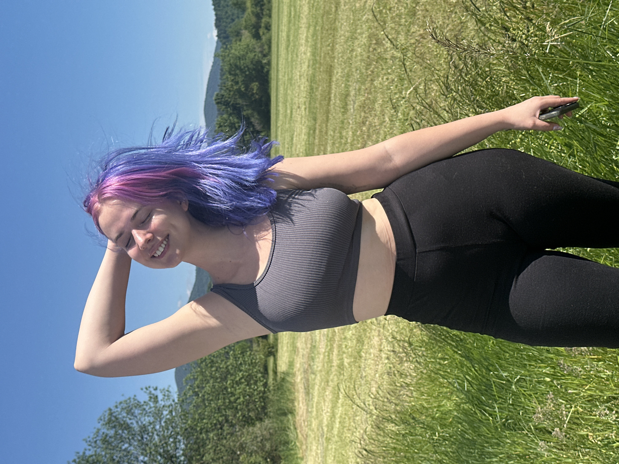 A young woman stands in a grassy field under a bright blue sky. She is wearing a grey ribbed crop top and black leggings. Her hair is dyed with pink and purple streaks. She is smiling and has one arm raised, touching her head. In her other hand, she holds a dark-colored smartphone. The background consists of a green field, trees, and distant mountains. The location is presumed to be near Postojna, Slovenia, due to the presence of karst landscapes.