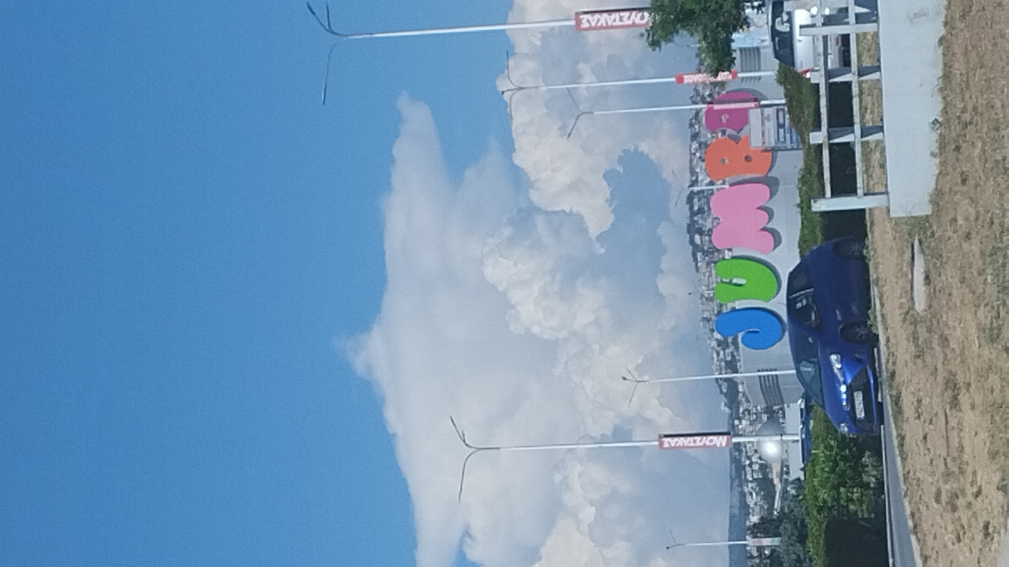 The image captures a bright, clear day in Pylaia Municipal Unit, Greece, with a striking blue sky dominating the upper portion of the frame. A prominent, large-scale sign displaying the word "JUMBO" in vibrant, distinct colors (blue, green, pink, orange, and purple) is visible. The sign is positioned in front of a building with a metallic facade.
In the foreground, a dark blue car is parked on the left side of the road, facing the sign. A portion of a silver car is visible on the right side of the frame, behind a white metal railing. The ground in the immediate foreground consists of dry, sparse vegetation and dirt.
Several tall streetlights with multiple arms are spaced along the roadway. Red and white banners are attached to two of the lampposts. In the background, a city skyline with buildings is spread across a hillside under a dramatic display of large, white cumulus clouds that create a sense of depth. Green foliage is visible on the left and right sides of the frame, partially obscuring the view of the surrounding area.