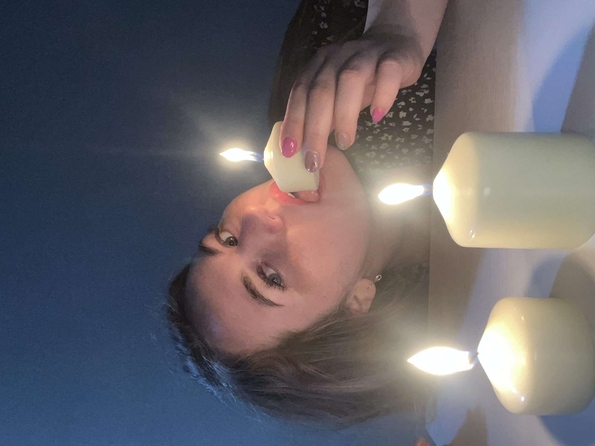 The image, rotated 90 degrees clockwise, presents a close-up, low-angle shot of a young person's face and upper body, appearing upside down. The lighting is dim, primarily emanating from three lit candles.
The subject is a young person with dark hair, green or hazel eyes, and what appears to be pink lipstick on their lips. They are wearing a dark garment with a light, possibly floral, pattern. Their left hand, adorned with alternating pink and iridescent nail polish, holds a small, hexagonal-shaped lit candle, bringing the flame very close to their open mouth and tongue. The person's gaze is directed upwards, towards the camera.
Two other larger, similarly shaped lit candles are visible to the right and bottom right of the frame, casting warm, glowing light across the scene. The background transitions from a dark blue on the left side to a lighter, possibly cream or white, wall on the right.
The scene appears to be set indoors during nighttime or in an artificially dimmed room, with the candles serving as the main light source. The activity suggests a deliberate, artistic pose or performance. There is no visible text in the image. The location context provided is Chaikovsky, Russia.