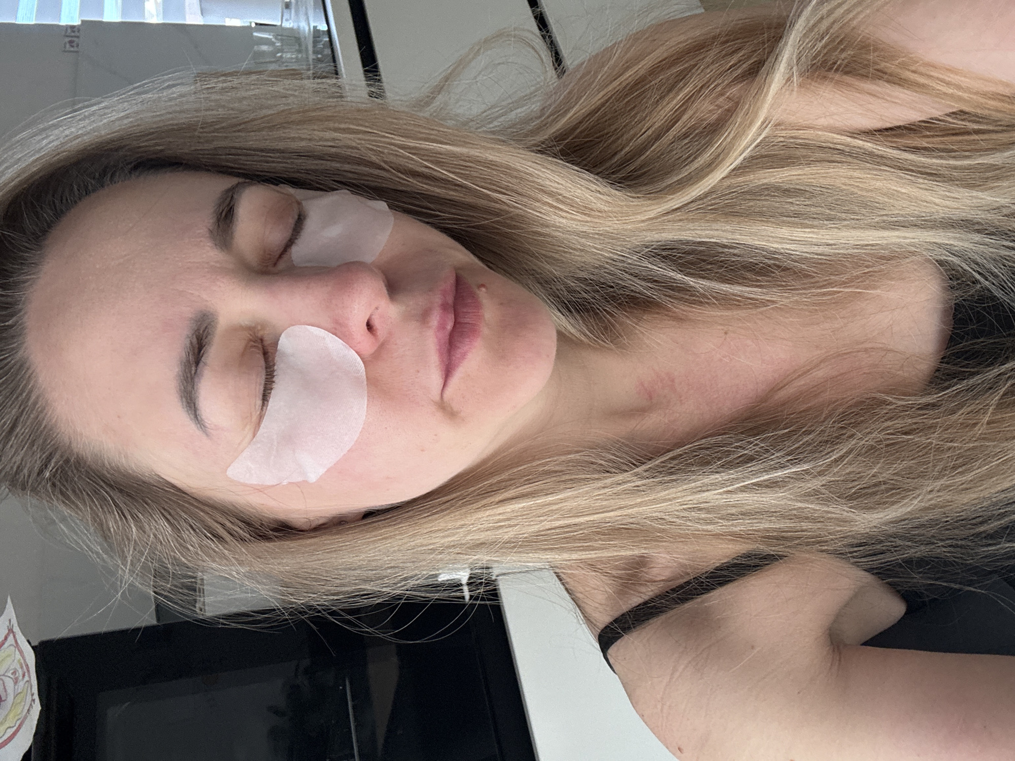 A woman with light-blonde hair is lying down with white cosmetic patches under her eyes. She appears to be in a domestic setting, possibly a kitchen, suggested by the presence of what seems to be a refrigerator in the background. The photograph is taken from above and slightly to the side. It does not include any specific details indicating the location is Chaikovsky, Russia.