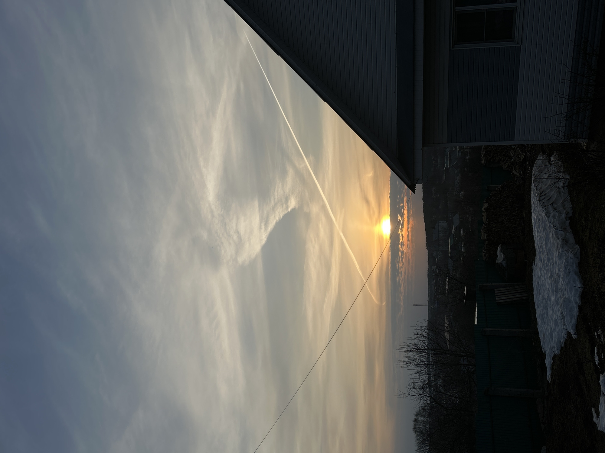The image shows a sunset scene in Фоки, Russia, viewed from what appears to be a residential area. The sun is setting on the horizon, creating a warm, orange glow in the sky. There are contrails from airplanes crisscrossing the sky. In the foreground, a portion of a building with siding is visible on the right side of the frame. Below the building, there is a fence, some trees, and patches of snow on the ground, indicating it is either winter or early spring. The lighting suggests the photo was taken around dusk.