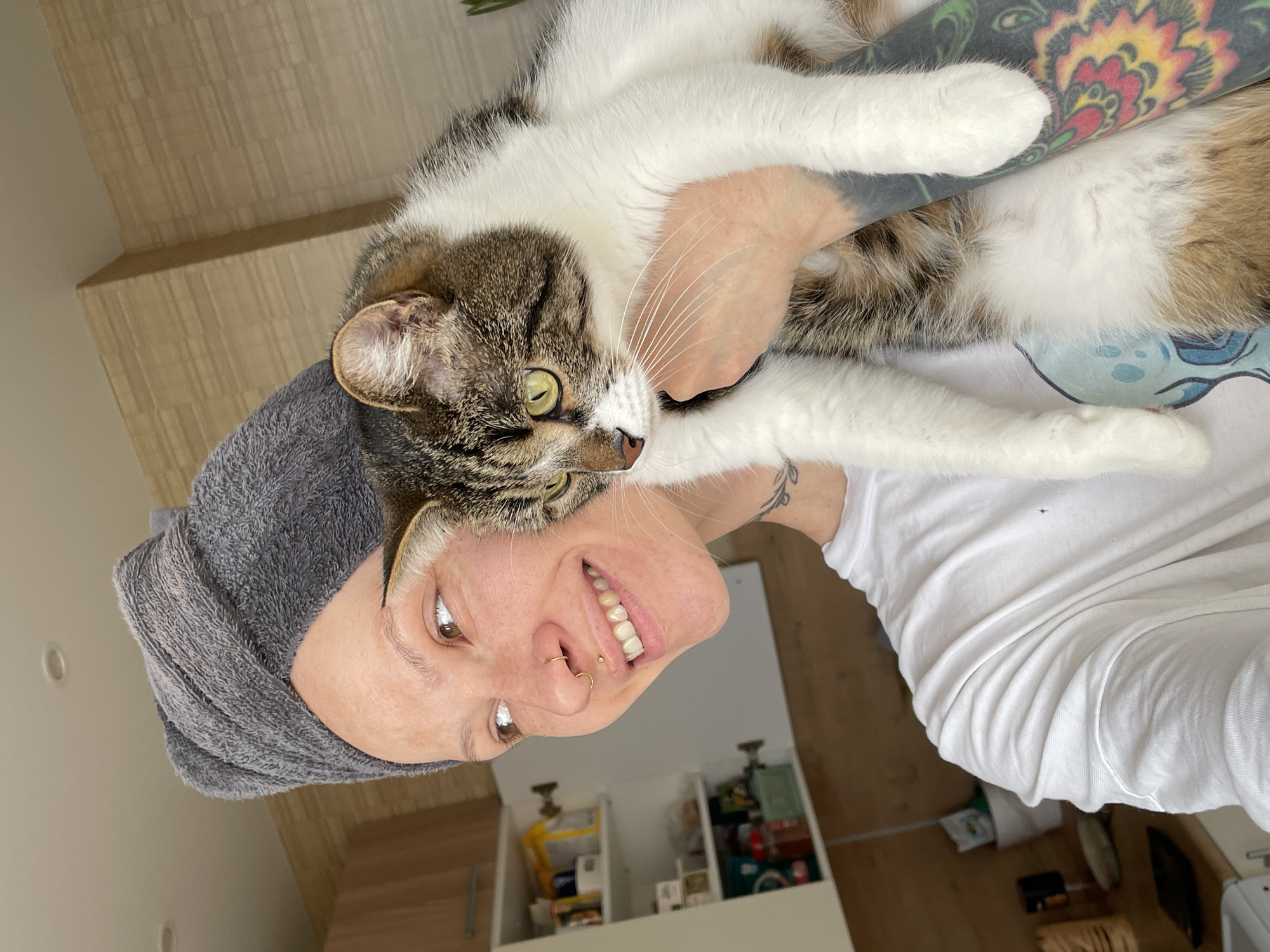 A person with a gray towel wrapped around their head is holding a cat. The cat is white and brown tabby. The person is smiling and has a nose piercing. The person has a tattoo on their arm. The background is a room with light-colored walls and wooden floors. There is a white cabinet in the background with items inside. It is not possible to determine the location in the city of Izhevsk, Russia from this image.