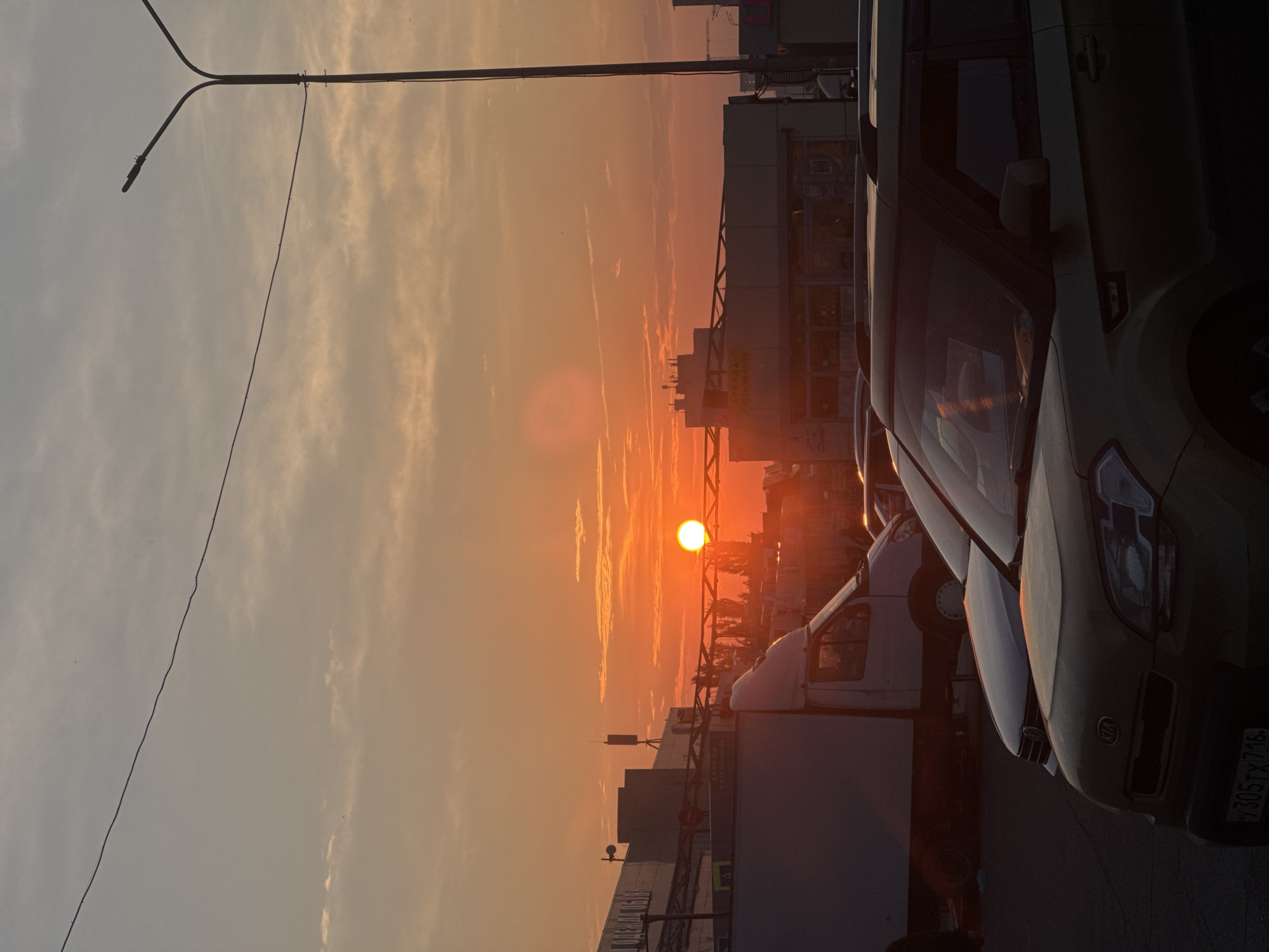 The image depicts a sunset in an urban environment, likely in Kazan, Russia. The sun, a bright orange orb, is low in the sky, casting a warm glow over the scene and illuminating thin, wispy clouds. In the foreground, several vehicles are parked, including a gold Kia Soul in the lower right and a white truck on the left. Beyond the vehicles, commercial buildings with flat roofs and signage are visible, silhouetted against the vibrant sky. A tall street lamp stands on the right side of the frame, its dark silhouette contrasting with the bright sunset. The overall atmosphere is one of a city at dusk, with the natural beauty of the sunset juxtaposed with the urban landscape.