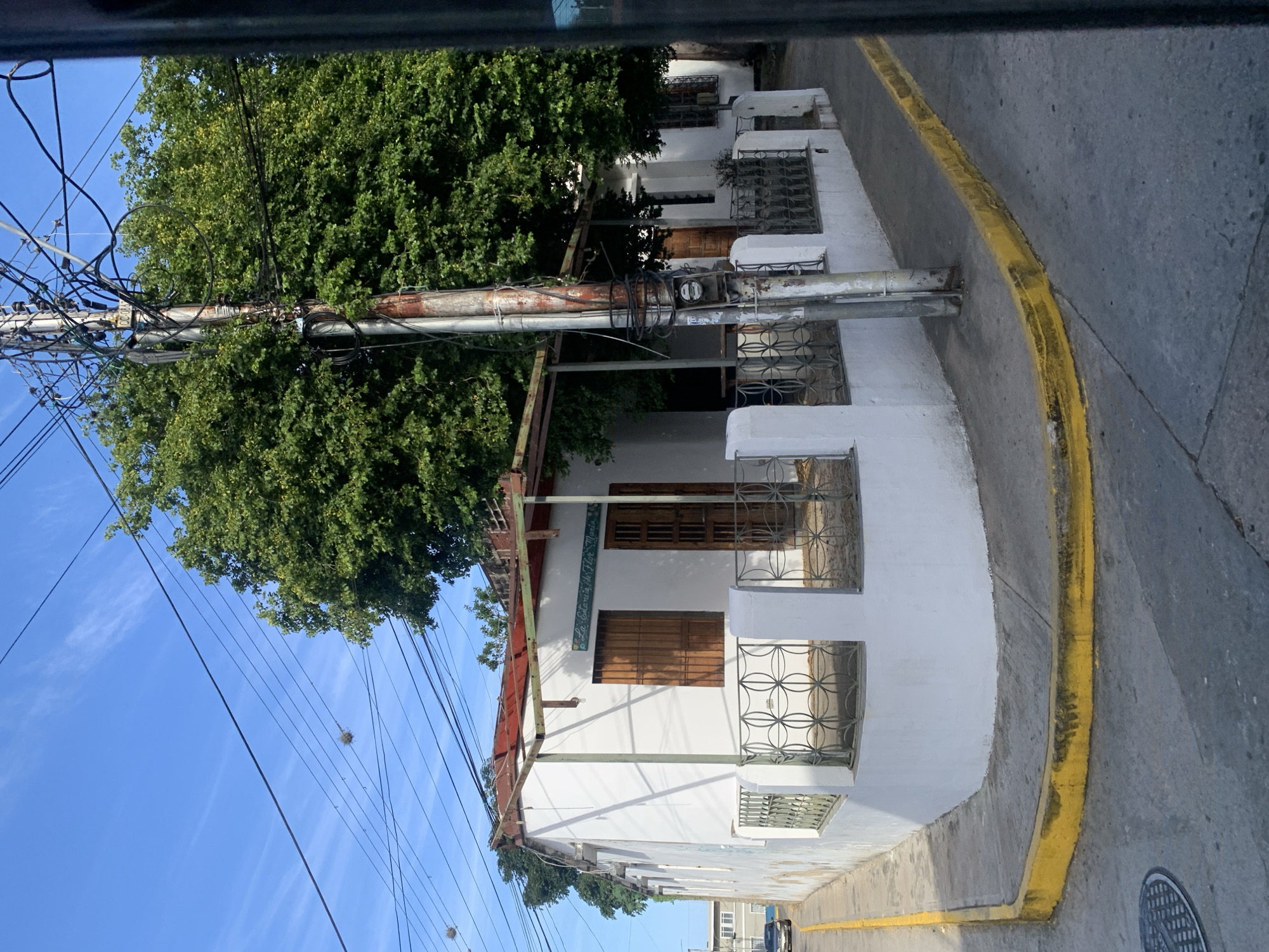 The image captures a street corner in La Asuncion, Venezuela, featuring a white, single-story building with a red-tiled roof and wooden shutters. The building appears to be a commercial establishment, indicated by a sign above the entrance that reads "La Estancia de Flor Maria". The property is partially enclosed by a low white wall with decorative metal fencing. A large, leafy tree shades a portion of the building. Several electrical poles and a complex network of overhead wires are prominent, a common sight in many urban areas. The street is paved with asphalt, and a yellow-painted curb marks the edge of the road. The sky is blue with wispy clouds, suggesting clear weather. A dark car is visible on the road in the background, suggesting normal urban activity. The overall scene depicts a typical streetscape in the historic city of La Asuncion.
