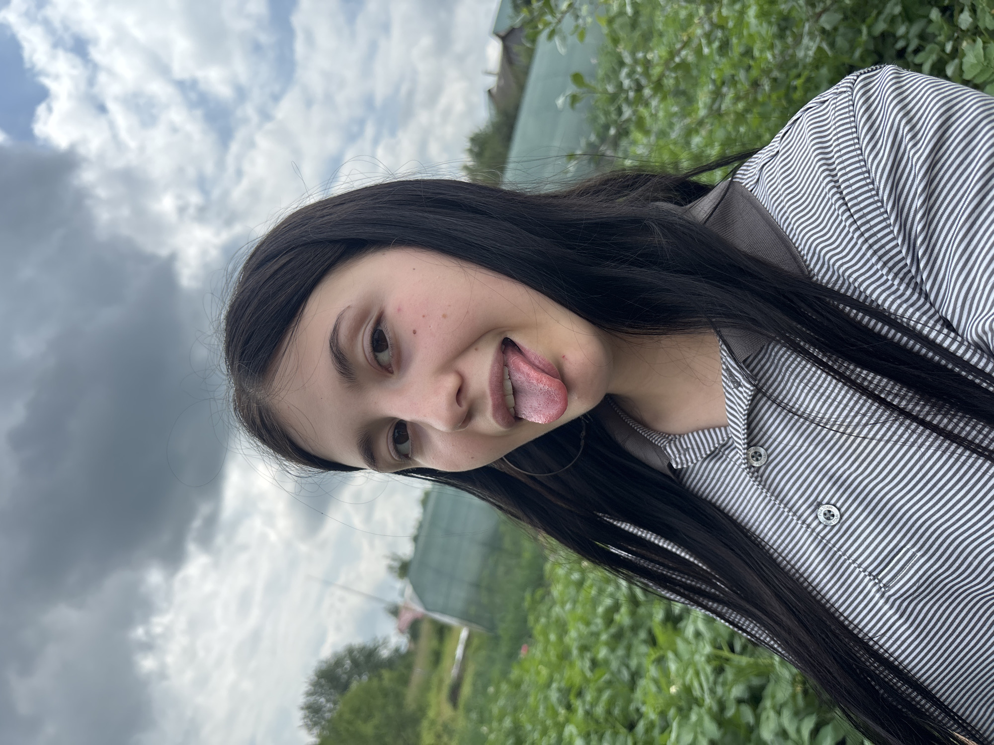 A young woman with long, dark hair is shown in a close-up, selfie-style shot outdoors. She is wearing a grey and white striped shirt. Her tongue is sticking out of her mouth. She has light-colored skin and a smattering of freckles. In the background, there are green plants, a greenhouse, and a cloudy sky. The location is described as the city of Fokino, Russia.