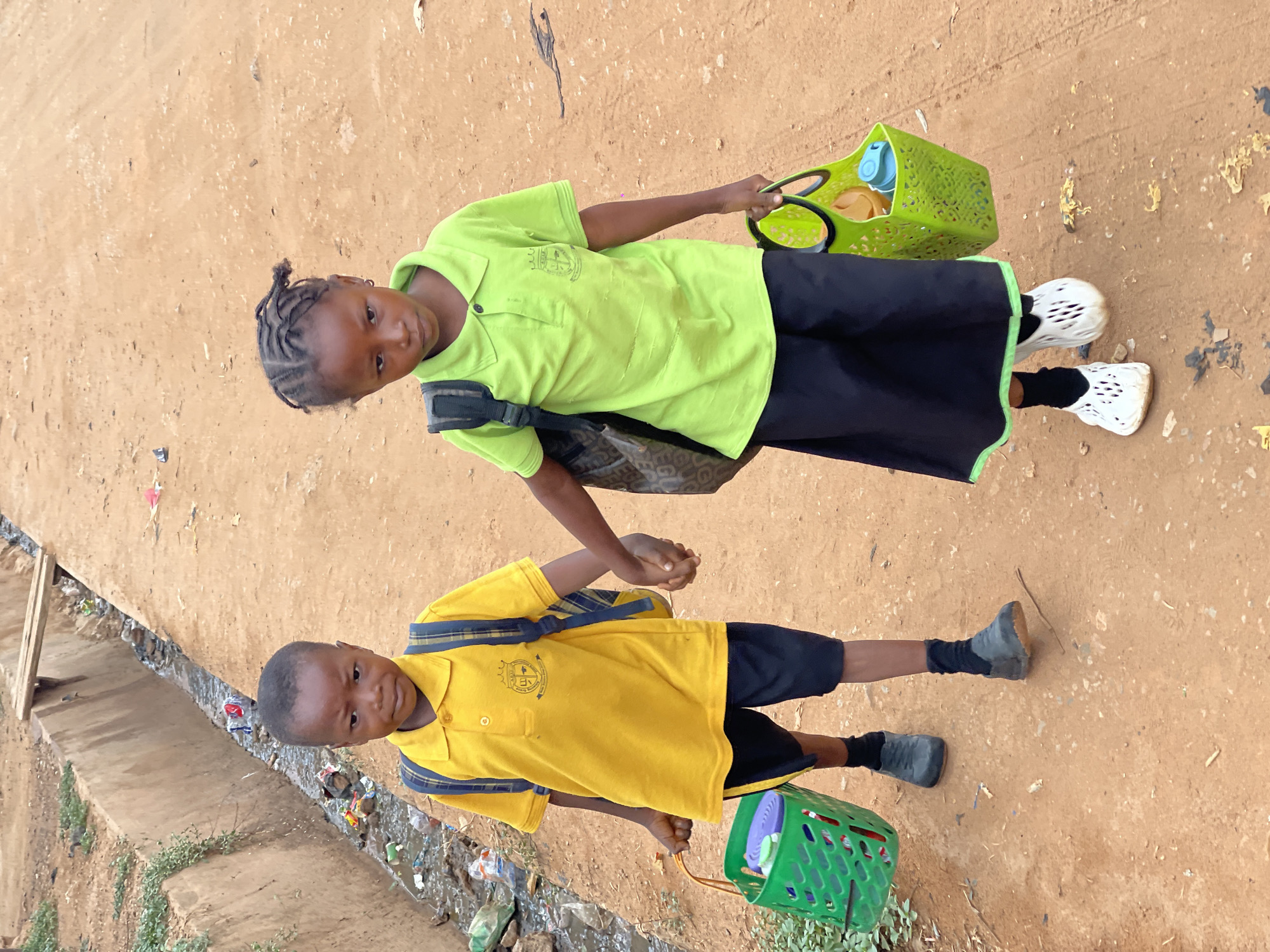 Two young children are depicted in the image. The child on the right wears a green short-sleeved polo shirt and a black skirt and carries a green basket. The child on the left wears a yellow short-sleeved polo shirt and black shorts, and also holds a green basket. They are holding hands. They appear to be standing on a dirt surface with what appears to be a drainage ditch filled with trash running along the side. This likely depicts an environment in Kuchingoro, Nigeria.
