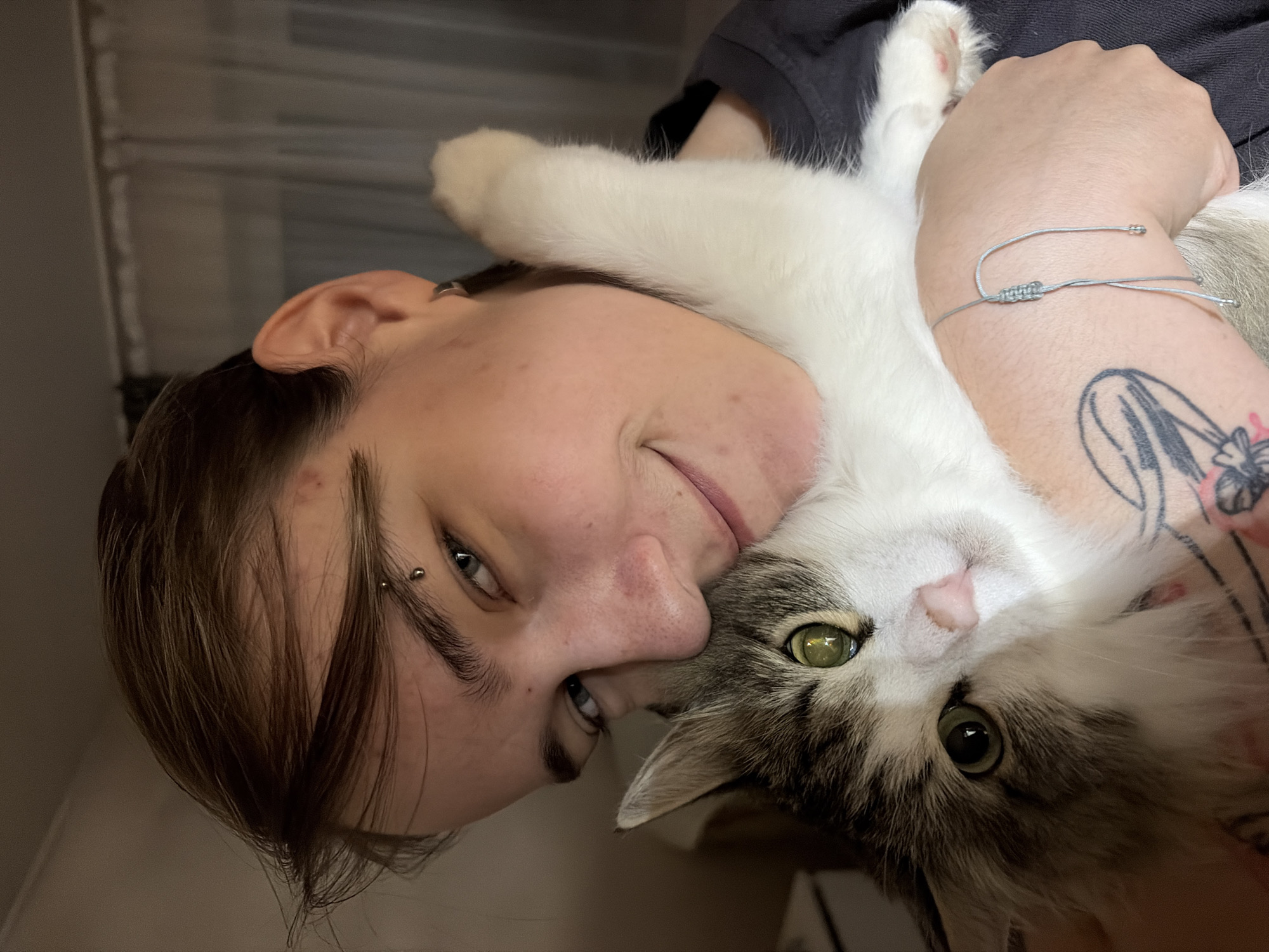 A person is holding a cat. The person has light skin, short brown hair, an eyebrow piercing, and a bracelet on their left wrist. A tattoo is also visible on the person's left arm. The cat has white and grey fur and green eyes. The photo appears to be taken indoors, possibly near a window or textured wall. The location in the city of Chaikovsky, Russia is not discernible from the visual information.