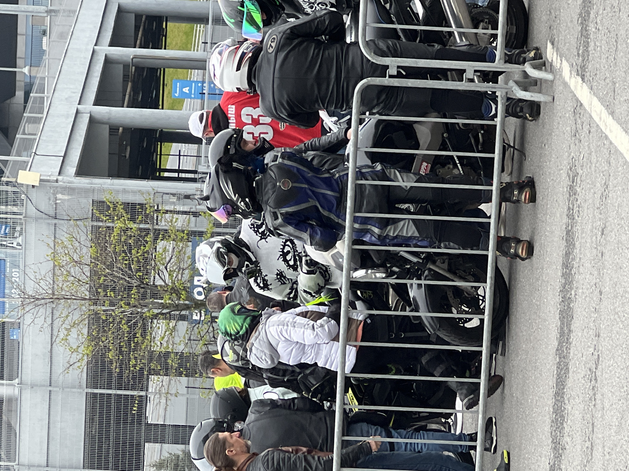 The image shows a group of motorcyclists, mostly wearing riding gear and helmets, standing behind a metal barrier. The motorcycles are parked closely together behind the barrier. In the background, there is a multi-story building with a metallic facade, typical of modern architecture. Signage in Russian is visible. Based on the architecture and signage, the location is likely in the city of Saint Petersburg, Russia.