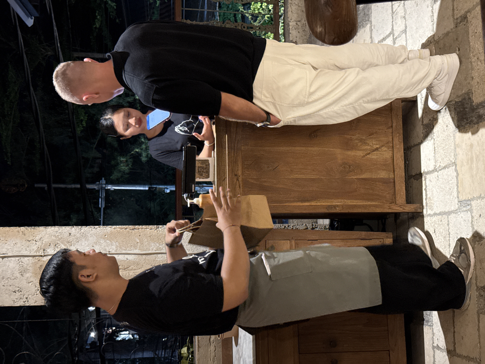 The image captures a nighttime scene at what appears to be an outdoor or semi-outdoor food or drink counter. The photo is rotated 90 degrees clockwise, so the description assumes a corrected orientation.
In the foreground, to the right, a man with short dark hair, wearing a black t-shirt and a grey apron, stands behind a wooden counter. He is looking slightly upwards and holding a brown paper bag with both hands, seemingly preparing or handling an order. A light-colored dispenser bottle is visible on the counter near his hands. He wears dark sneakers.
Opposite him, on the other side of the counter, is another man with very short blonde hair. He is wearing a black long-sleeved shirt, light-colored (off-white or beige) pants, and white sneakers. He is leaning slightly towards the counter, appearing to be a customer interacting with the server. A watch is visible on his left wrist.
In the mid-ground, behind the blonde-haired man, a woman with dark hair is visible. She wears a dark t-shirt and is looking intently at an illuminated smartphone screen.
The background is dark, indicating night, with hints of greenery and some indistinct structures. The ground is paved with light-colored stones or bricks. The overall activity depicts a transaction or interaction at a service counter, possibly at a cafe or street food stall. No visible text is clearly discernible.