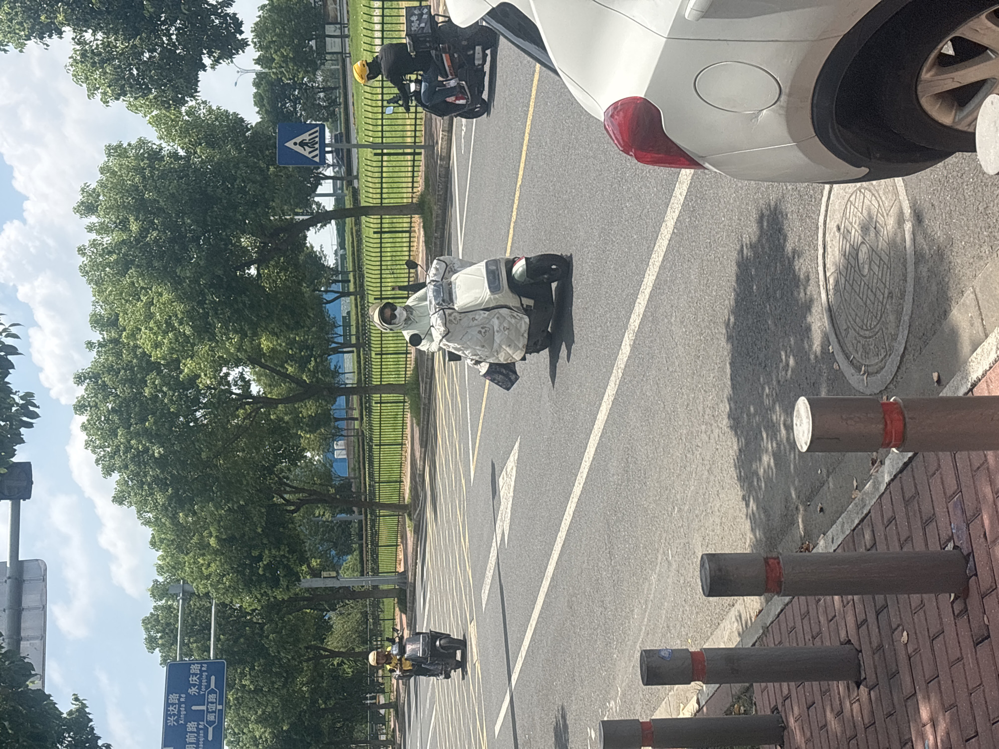 In Suzhou Industrial Park, China, on a sunny day with a cloudy sky, three individuals on scooters navigate a street. The foreground is dominated by the rear of a white car, its taillight red and a fuel cap visible on the rear quarter panel. The street is paved with asphalt and marked with white lines, including directional arrows. On the right side of the road, several bollards with red bands line a red brick sidewalk.
The first scooter, ridden by a person wearing a yellow helmet and a yellow top, is in the middle lane, moving away from the viewer. The second scooter, a white one, is in the same lane, closer to the center. The rider is wearing a light-colored hooded jacket, sunglasses, and a white face mask, with a large, patterned fabric cover draped over the front of the scooter. The third scooter, further back in the right lane, is ridden by a person in a yellow helmet and a dark top, with a black box mounted on the rear.
In the background, a green metal fence separates the road from a grassy area with trees. A pedestrian crossing sign featuring a blue background with a white figure is visible further down the road. Above the fence and trees, a blue road sign with white text indicates directions, showing "Xingda Rd" and "Tongqing Rd." The overall scene depicts a typical urban streetscape with moderate traffic.