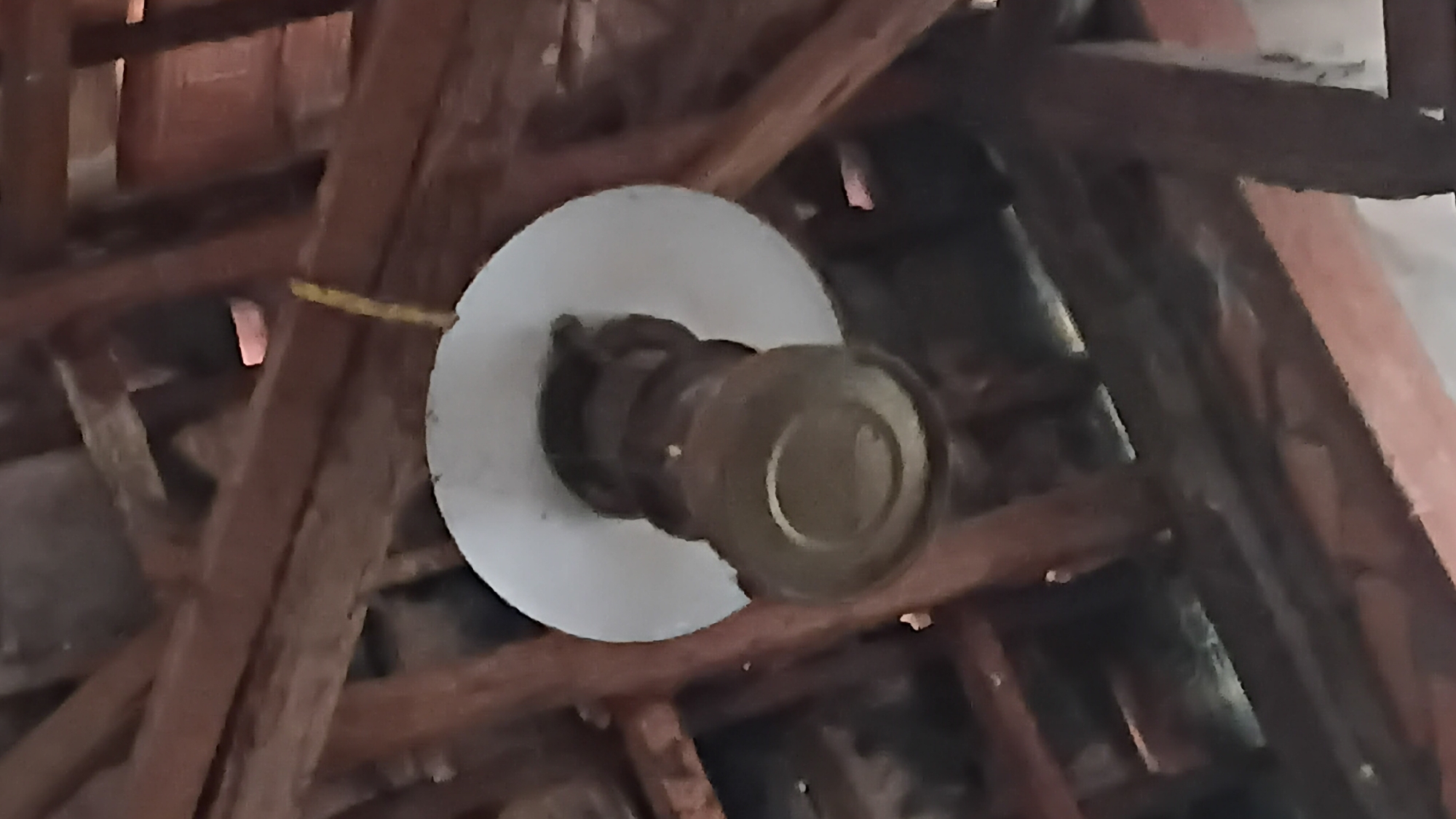 The image shows a light fixture hanging from wooden beams, likely in an attic or under a roof. The light fixture has a white disc-shaped shade and a brown, cylindrical body with several stacked sections. A yellow string is attached to the fixture and hangs down. The wooden beams are dark brown and appear to be rough-hewn. There is some debris visible between the beams. The scene suggests an interior space with exposed wooden structural elements. No people are visible in the image, and there are no clear indications of the specific location within Prabumulih, Indonesia, beyond the general context of a building's structure.