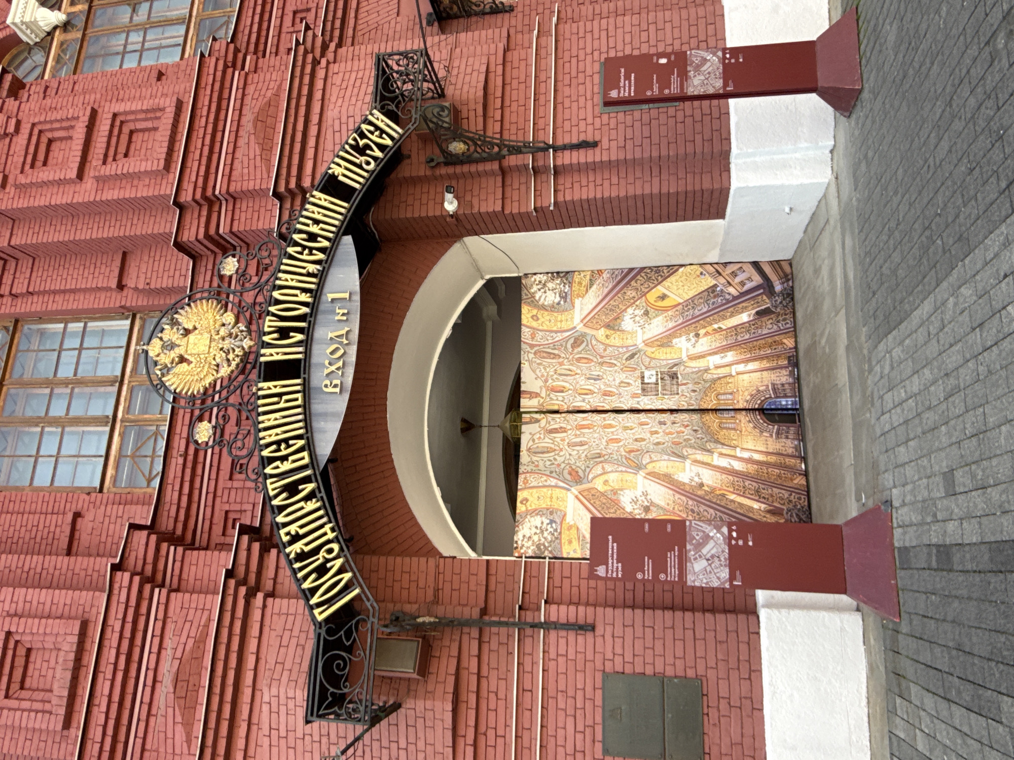 This image captures the entrance of the State Historical Museum in Moscow, Russia. The museum's red brick facade is prominent, showcasing ornate architectural details. An arched entrance is adorned with a black metal sign in Cyrillic, with gold lettering denoting the museum's name. Inside the entrance, the doors feature intricate, colorful patterns. To the sides of the entrance are information signs, presented in maroon. The image is slightly tilted, giving a dynamic perspective of the building. The foreground includes a section of grey paved ground.