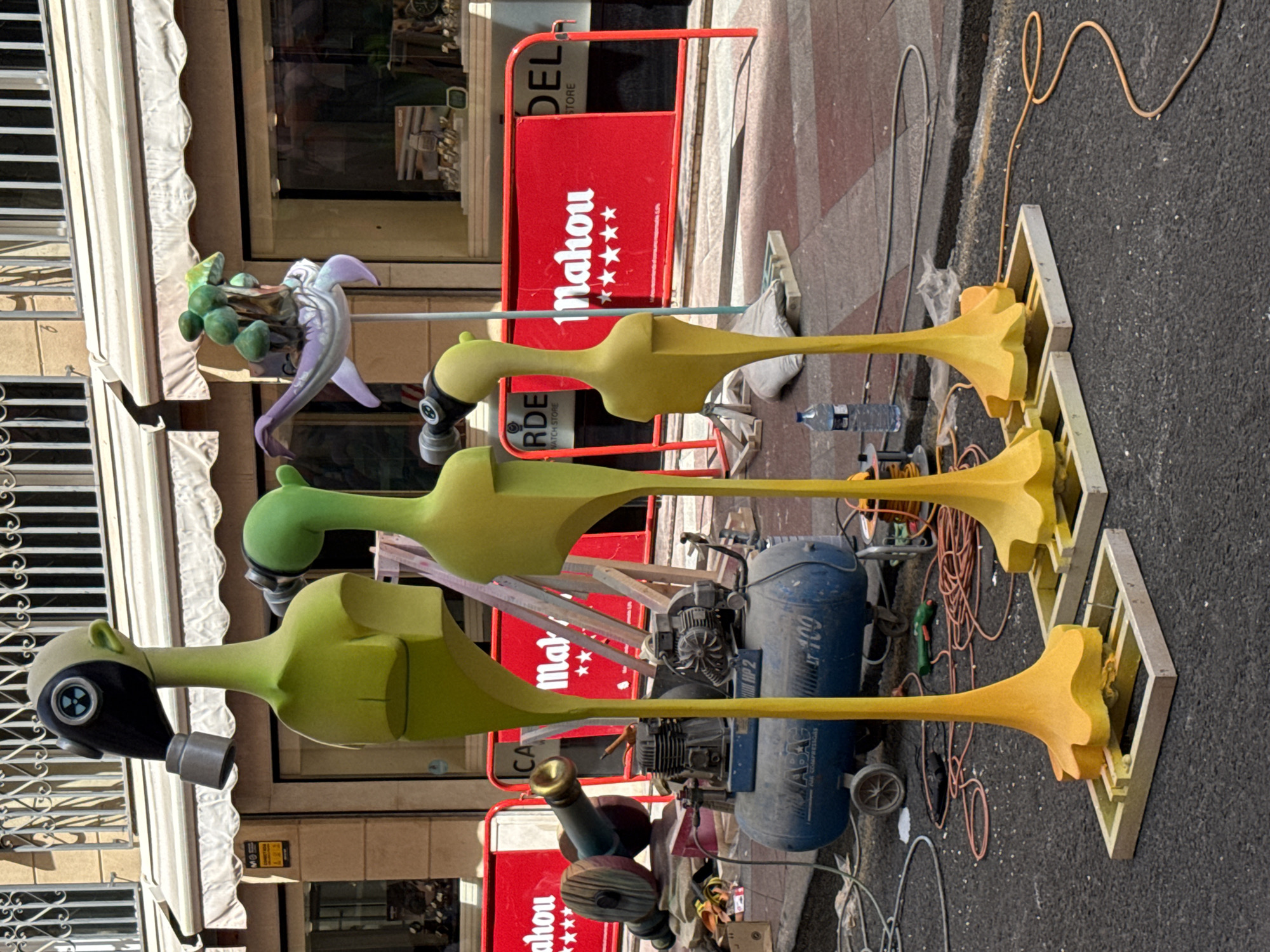 The image shows an outdoor scene in an urban setting, likely a street, with the facades of buildings visible in the background. The primary subjects are three tall, slender, anthropomorphic figures, painted a bright yellow-green. Each figure has a stylized head resembling a bird or an alien, and one of the figures is wearing a black gas mask with a circular filter. The figures are standing on wooden bases.
In the mid-ground, there are several objects and structures that suggest ongoing work or preparation. A large, blue air compressor sits on wheels. Red barricades with white lettering ("Mahou") are positioned across the street. A cannon-like object with wooden wheels and a brass muzzle lies on its side behind one of the barricades. Electrical cords and hoses are scattered on the asphalt. A bottle of water is visible near one of the figures. A purple and green sculpture of a creature with mushrooms on its back is mounted on a pole behind the figures. The ground surface is asphalt, with some cobblestone or tiled areas visible in the background. The lighting suggests it is daytime, with shadows cast by the figures and objects. The overall impression is a street scene during an event setup or breakdown.