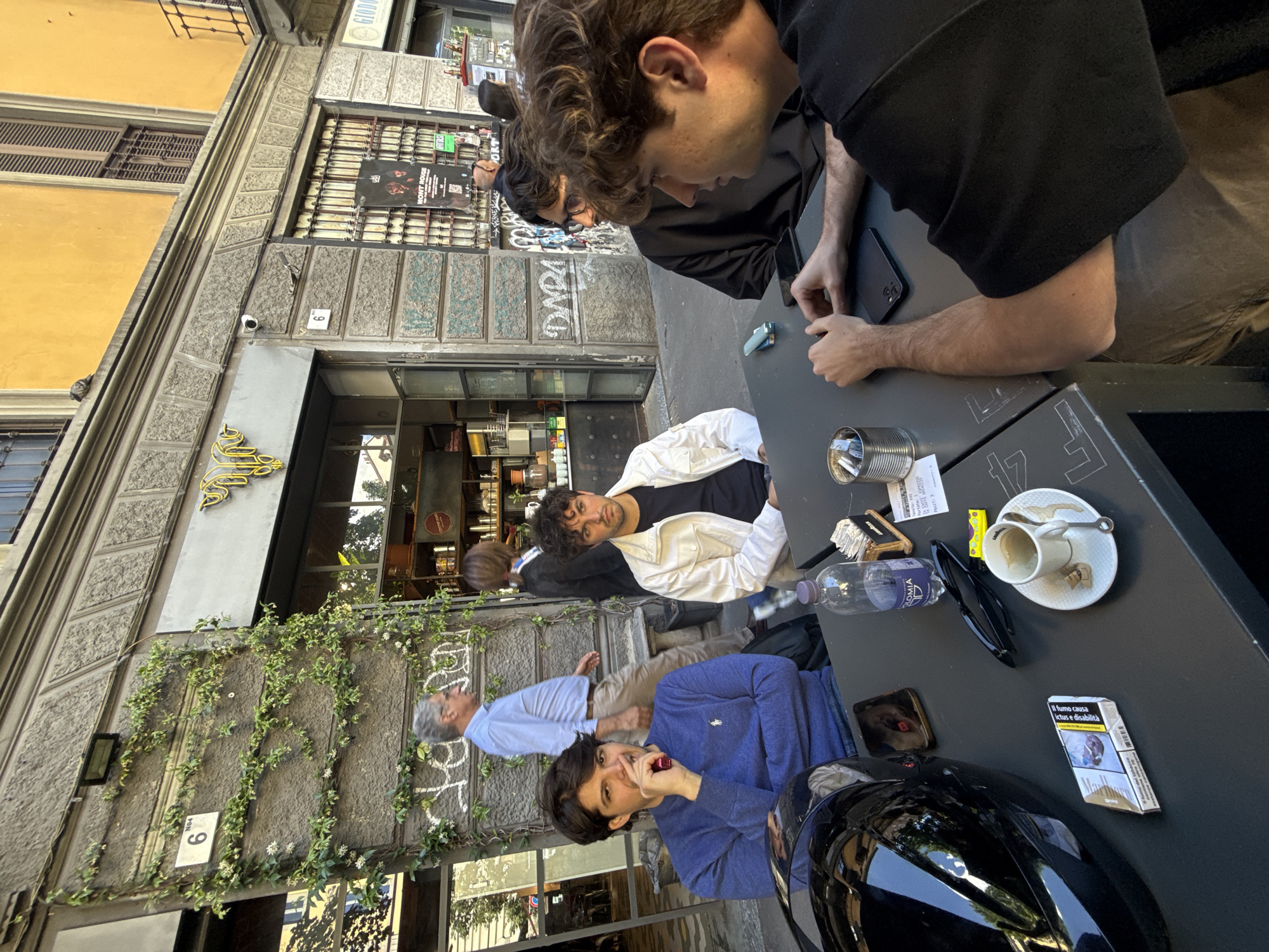 This photo, taken outdoors, shows four men at a table outside a shop in Milan, Italy. The shop is marked with the number "6" and the word "GIODO" above the entrance. The exterior of the building has light-colored walls with an ivy-covered portion.
Three of the men are seated at a black table. The man closest to the camera is wearing a blue shirt and is holding a small object to his mouth. A black motorcycle helmet is next to him on the table. Next to him is a man in a white jacket over a black shirt. The third man at the table is wearing a black shirt and is looking down at a phone or another small object on the table. The table is covered with a water bottle, a pack of cigarettes, a pair of sunglasses, a small yellow container, and an empty coffee cup. An older man in a light blue shirt is standing in the background. Graffiti is visible on the wall.