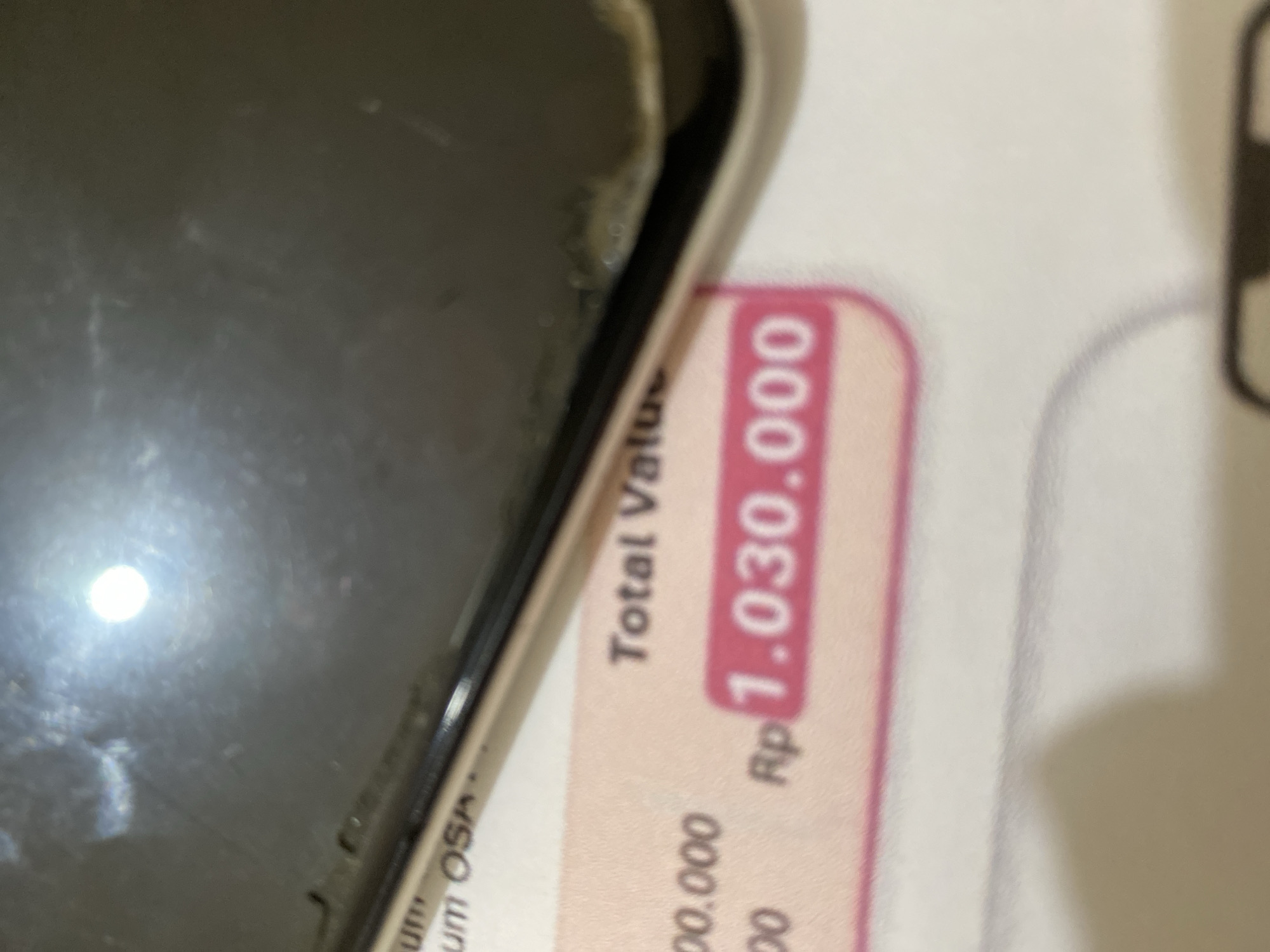 The image shows a close-up of a portion of a phone's screen placed over a document. The document appears to be a receipt or invoice with text and numbers printed on it. Prominently visible is the text "Total Valu" followed by "Rp 1.030.000". Other partial text like "um OSA" and "00.000" are also visible on the document. The phone's screen has a bright reflection on it, obscuring some of the details. The background is white and out of focus. There is no discernible information about the city of Lhokseumawe, Indonesia, in the provided image.