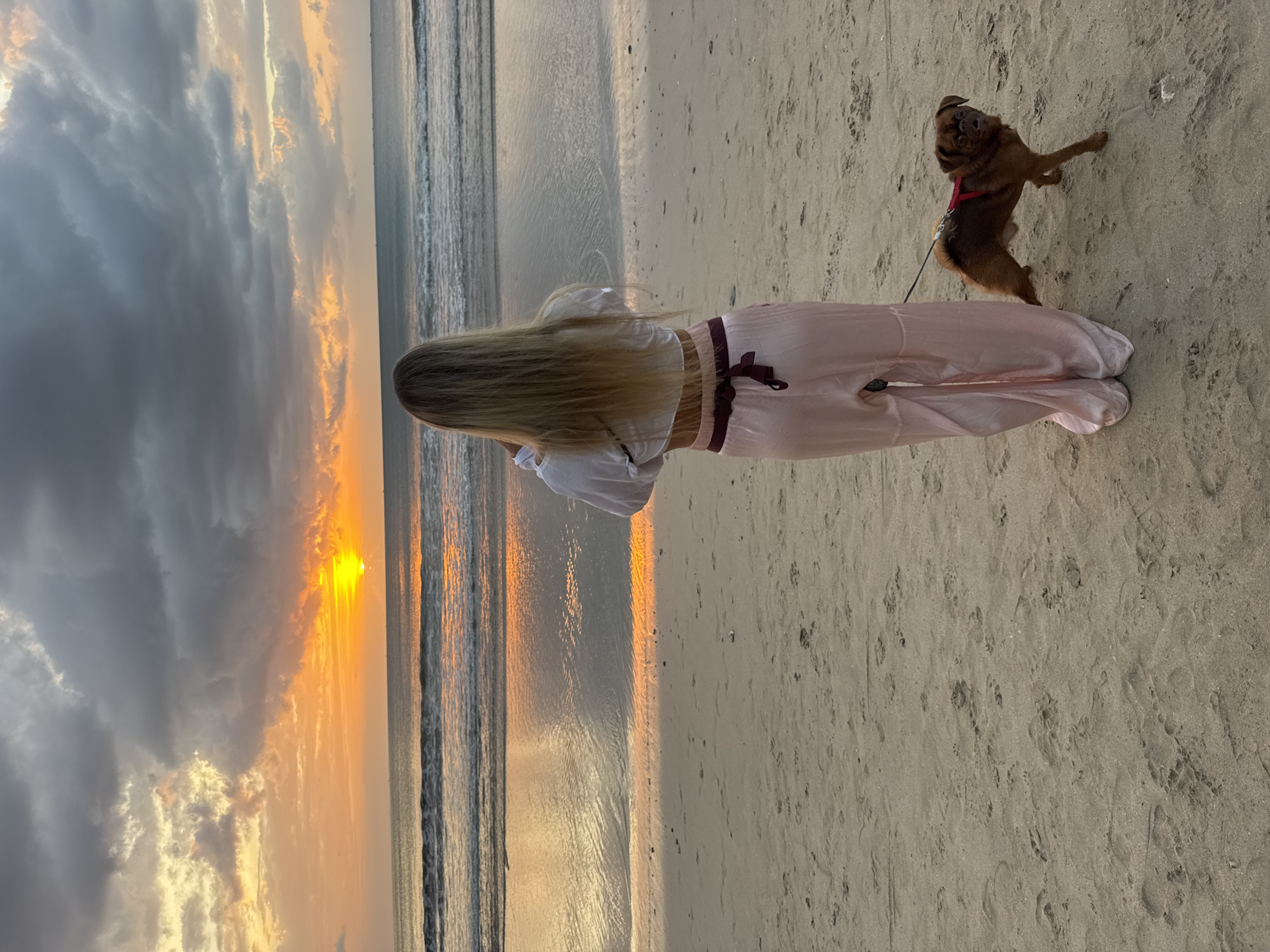 A person stands on a sandy beach, facing away from the camera, looking out at the ocean during sunset. The sky is filled with dramatic clouds, ranging from dark grey to vibrant orange and yellow as the sun sets near the horizon. The ocean has gentle waves, and the wet sand reflects the colors of the sky. A small, brown dog on a leash stands next to the person. The person is wearing a white top and loose pink pants with a dark red sash. The beach is sparsely populated, with some footprints visible in the sand. The image appears to have been taken in Kuta Selatan, Indonesia, given the characteristic sunset and beach environment.