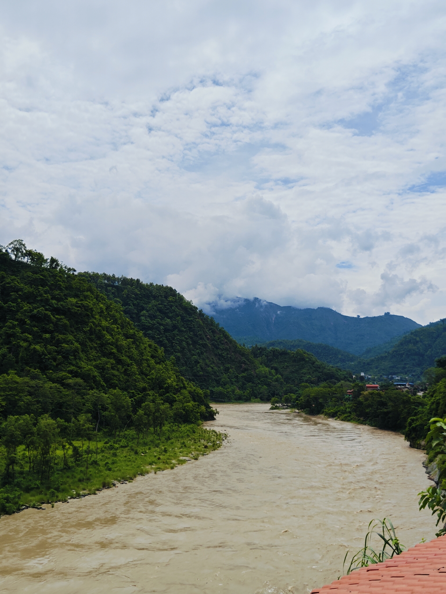The image depicts a wide, muddy river flowing through a valley in Ichchhakamana, Nepal. Lush green hills covered in dense vegetation rise steeply on either side of the river. In the distance, more mountains are visible, some shrouded in clouds. A small settlement with buildings can be seen nestled in the valley on the right side of the river. The sky is overcast with scattered white clouds. The foreground features a portion of a red-tiled roof and some green foliage. The overall scene conveys a sense of natural beauty and the power of the river in a mountainous region.