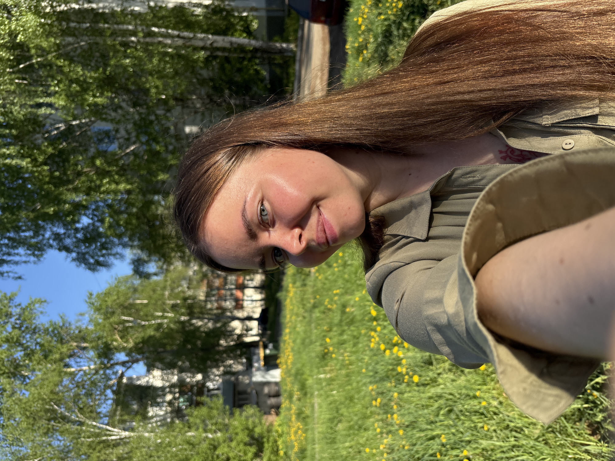 The image shows a woman with long, straight brown hair, wearing a green shirt. She is standing in a grassy area with yellow flowers, possibly dandelions. Behind her are birch trees and what appears to be a residential building. The location is most likely somewhere in Izhevsk, Russia, given the trees and building style are common to the area.