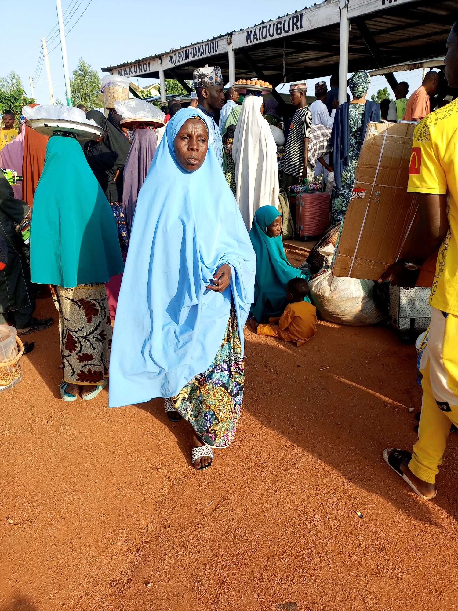 The image depicts a busy outdoor scene at what appears to be a transportation hub or market in Gombe, Nigeria. A group of people, primarily women wearing headscarves and traditional attire, are present, along with some men. The individuals are carrying or arranging various goods and luggage, suggesting a moment of transit or trade.
In the foreground, a woman wearing a bright blue headscarf and a patterned skirt is looking towards the camera. To her left, another woman in a turquoise headscarf and a similarly patterned skirt is carrying a tray on her head. Other individuals are visible in the background, some are carrying bags, boxes and other items. Some of the people are standing, while one woman is seated on the ground with a child in an orange outfit.
Overhead, a structure with signs displaying "MAKURDI" and "PATISKUM-DAMATURU" and "MAIDUGURI" suggests this location is a major route connecting these cities. The ground is covered in reddish-brown dirt, and there are some items scattered on it. The overall atmosphere appears to be dynamic and functional, indicative of daily life and commerce in the city. The lighting suggests it is daytime, possibly late afternoon, with shadows cast by the structure and people.