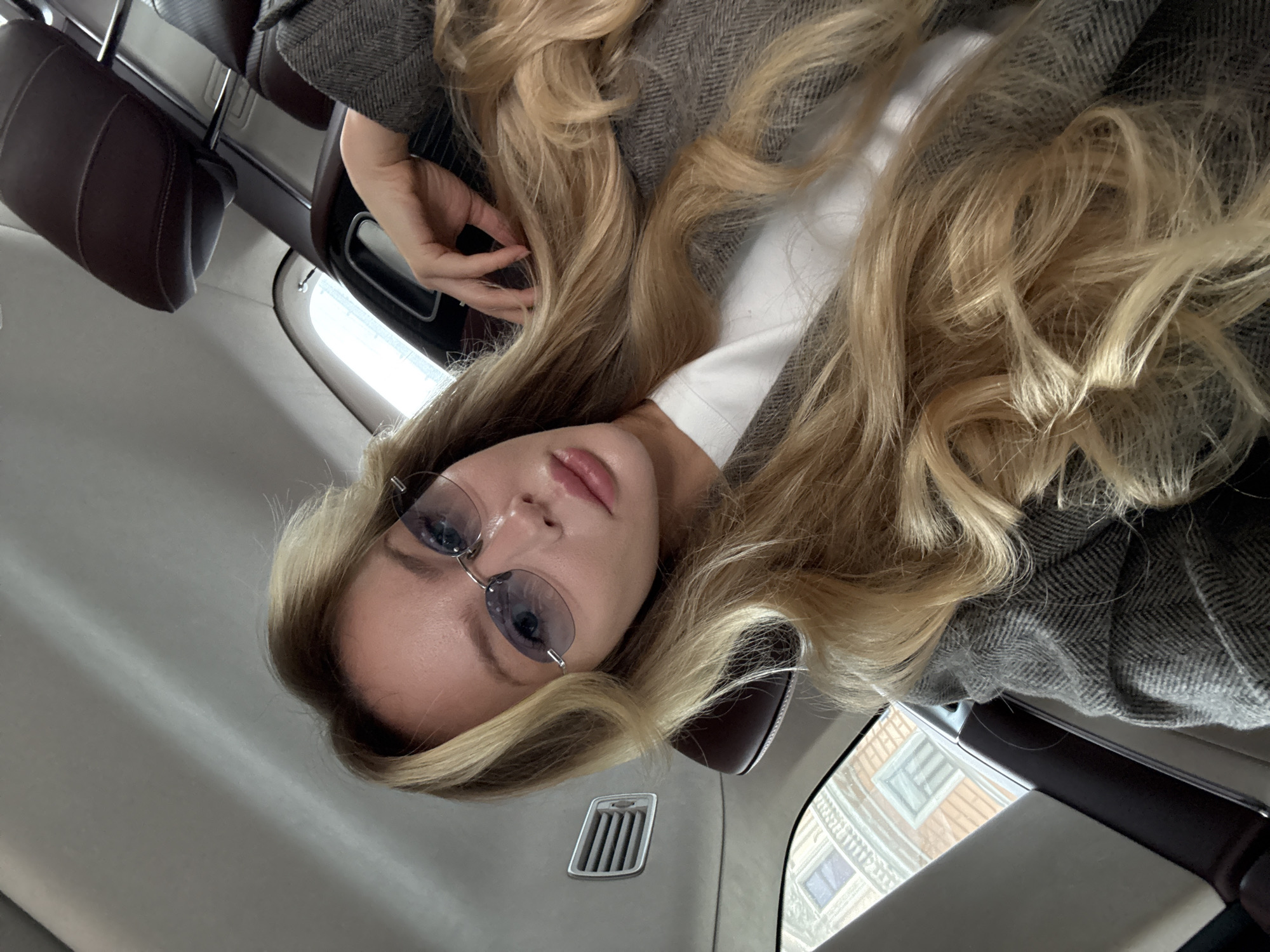 The image captures a young woman with long, wavy blonde hair, looking directly at the camera. She wears light-blue tinted, rimless glasses and has a neutral expression with slightly parted, pink lips. Her attire includes a grey jacket with a herringbone pattern over a white top. One of her hands is visible near her hair on the left side of the frame.
The setting appears to be the interior of a vehicle, such as a car or a train. Dark brown leather headrests and seat upholstery are visible, suggesting comfortable seating. The ceiling or upper interior panel is a light neutral color, featuring a silver-colored air vent. Through a window on the right side of the frame, a blurred urban building with visible windows can be seen, consistent with a city environment like Moscow. The lighting is soft and even, suggesting daytime conditions. There is no visible text in the image.