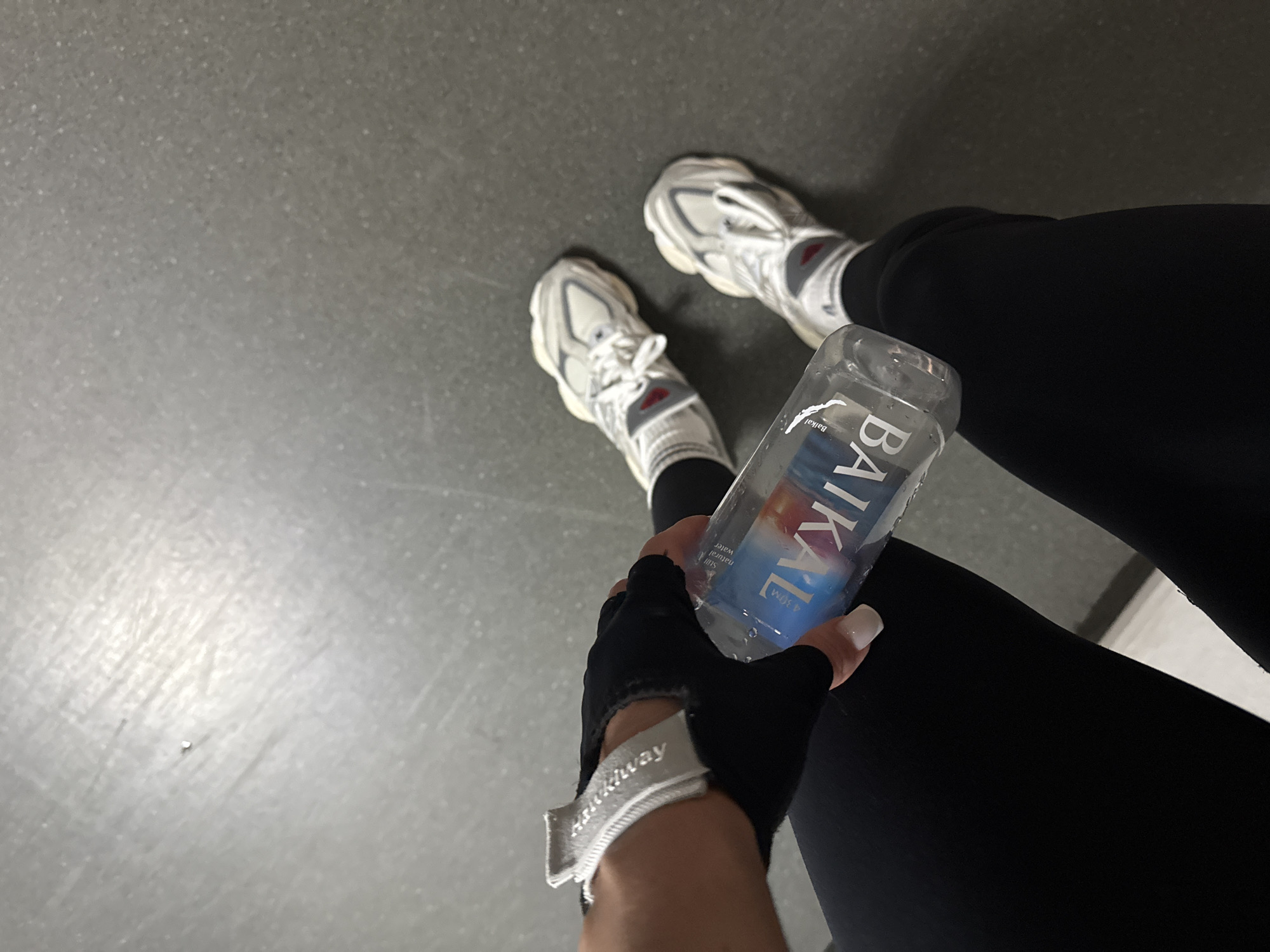 A person in black athletic leggings and white sneakers stands on a gray floor, holding a bottle of "Baikal" natural mineral water in a black athletic glove. The scene is likely indoors, possibly a gym or workout area. The location in Kazan, Russia is assumed due to the presence of the "Baikal" water bottle.