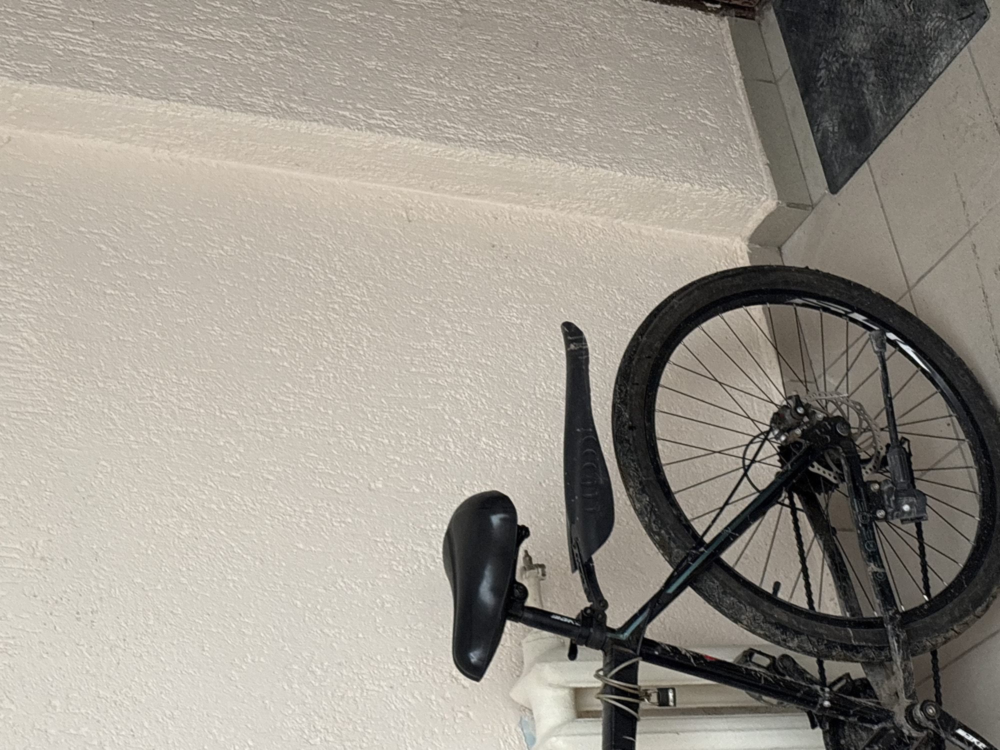 A bicycle is leaning against a wall. The bicycle is black and has a black seat, a black rear fender, and a black tire. The wall is light beige with a textured surface. A section of tiled wall in grays can be seen near the top right corner of the image. There is no evidence to verify that the image was taken in the city of Chaikovsky, Russia.