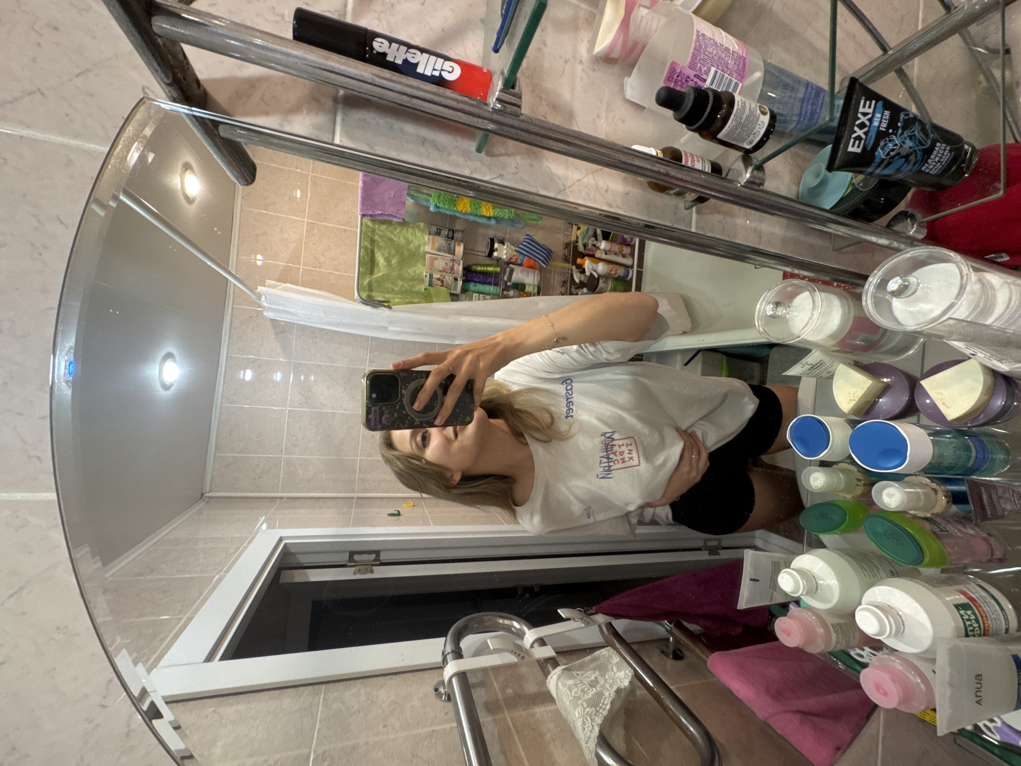 The image shows a selfie taken in a bathroom, likely in a residential setting, by a person reflected in a mirror. The person, presumably female, is holding a smartphone and wearing a white long-sleeved shirt and black shorts. The bathroom features tiled walls and a vanity area with visible toiletries and personal care products organized on a shelf above the sink. Brands like Gillette and Exxe are visible. A towel rack with towels hangs beneath the vanity. The image does not provide any information that would identify a specific location in Izhevsk, Russia, or any other location.