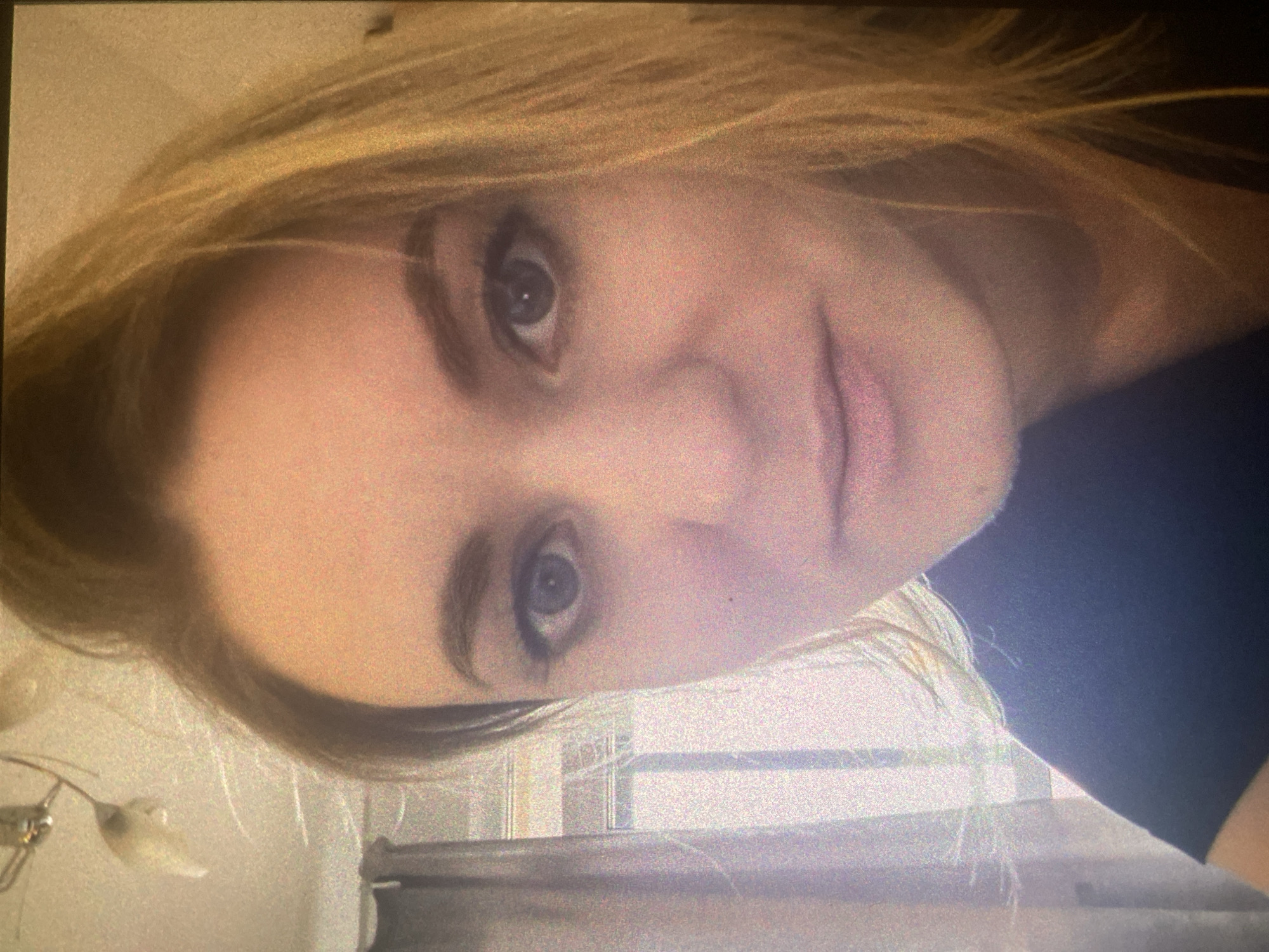 A portrait-oriented image, captured sideways, features a young woman with blonde hair and light-colored eyes, looking directly at the camera with a subtle, neutral expression. Her face is in a close-up, revealing fair skin. She wears a dark-colored top, visible on her shoulder and upper arm.
The background indicates an indoor setting, likely a room in Saint Petersburg, Russia. To what would be her upper-left (when viewed correctly), a light-colored wall is visible with a small, light-toned hanging object, possibly a light fixture or decoration. The lower and right parts of the background are blurred but show vertical and horizontal structural lines, suggesting a window with blinds or a piece of furniture. The overall lighting is soft, and the image exhibits some graininess, characteristic of indoor photography, possibly in lower light. No text is visible in the frame.