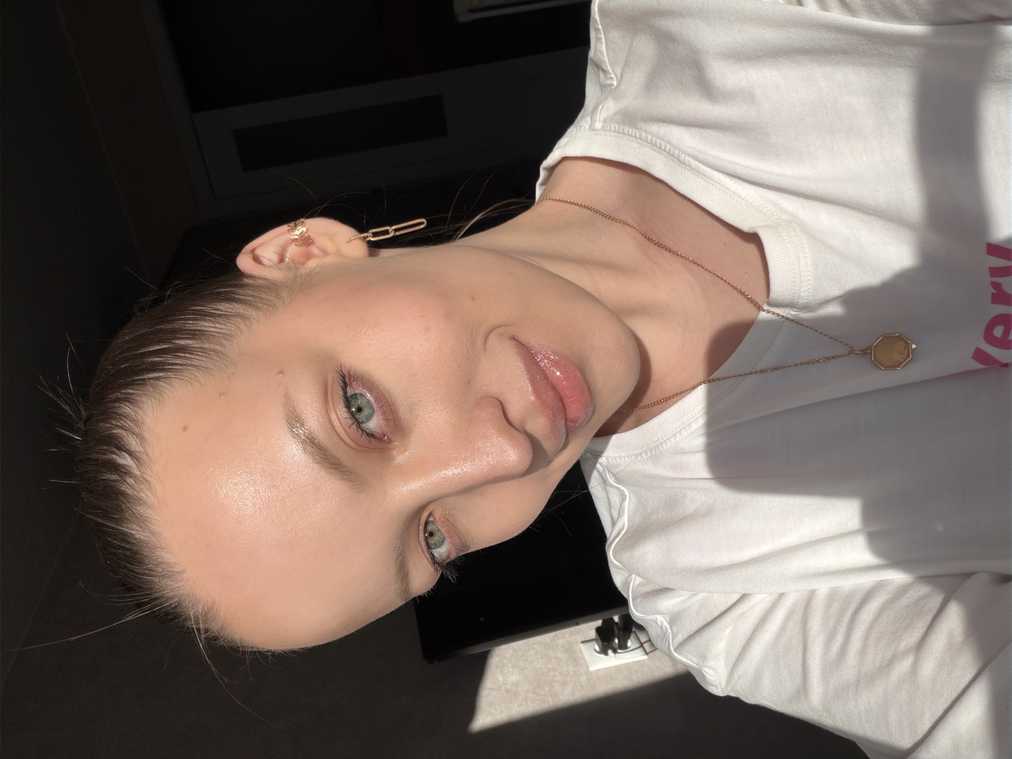 A female subject, an adult, is depicted in a close-up selfie within an indoor setting in Reshetnikovo, Russia. Her dark brown hair is pulled back, revealing a clear complexion and green eyes. She exhibits a slight smile and direct gaze towards the camera. She wears a white t-shirt, a portion of a pink graphic visible on the lower right. Adorning her right ear are a gold-colored leaf-shaped ear cuff on the upper cartilage and a dangling gold-colored earring with two linked oval shapes in the lobe. Around her neck is a thin gold-colored chain with an octagonal, gold-colored locket-style pendant. Strong, directional sunlight illuminates the left side of her face and body, creating sharp shadows. The background includes a light-colored, textured wall on the left with a white electrical outlet containing a dark plug, and largely indistinct dark, shadowed areas on the right.
