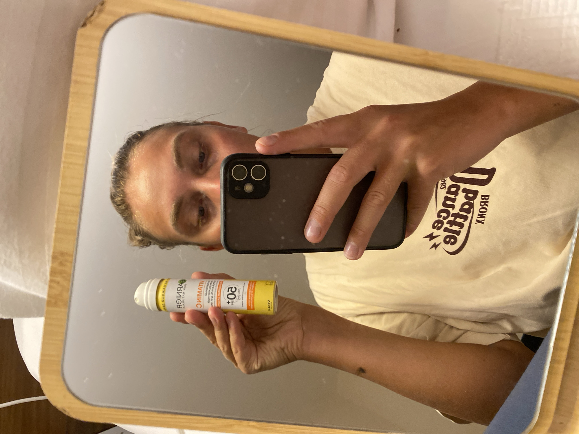 A person is taking a mirror selfie in a bathroom. They are holding a bottle of Garnier Vitamin C serum with SPF 50 in their left hand and a smartphone in their right hand. The person is wearing a cream-colored t-shirt with a graphic design. The mirror has a wooden frame. The background is out of focus but appears to be a tiled wall and ceiling. The person's face is visible in the mirror, looking at the phone's camera. The image does not provide any information about the city of Aksu, Turkey.