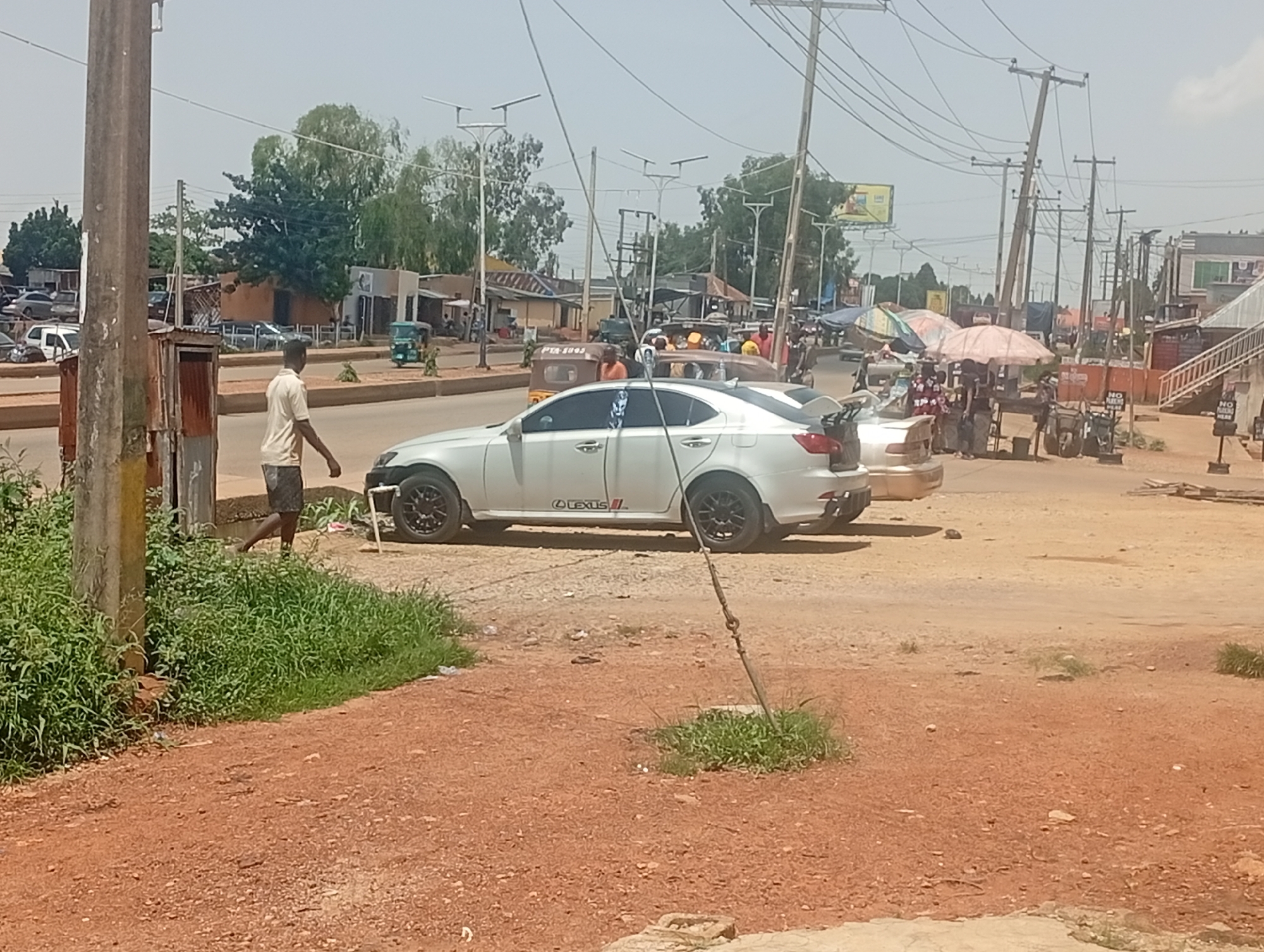 The image depicts a street scene in Bukuru, Nigeria, under a clear, bright sky, suggesting midday. A man wearing a light-colored short-sleeved shirt and dark shorts walks along the side of a dirt road. He is positioned between a utility pole and a white Lexus sedan, which has a spoiler and black rims. Lush green grass grows in patches along the roadside.
The background reveals a bustling environment with various vehicles and structures. A three-wheeled vehicle, commonly known as a "Keke Napep," is visible, along with other cars. Several market stalls are set up with umbrellas providing shade for vendors and their goods. Power lines crisscross the sky, supported by numerous utility poles. Buildings, some appearing to be shops or homes, line the street. The overall impression is one of a busy commercial area. The ground is a mix of red earth and scattered debris. The lighting and the clear sky indicate fair weather.