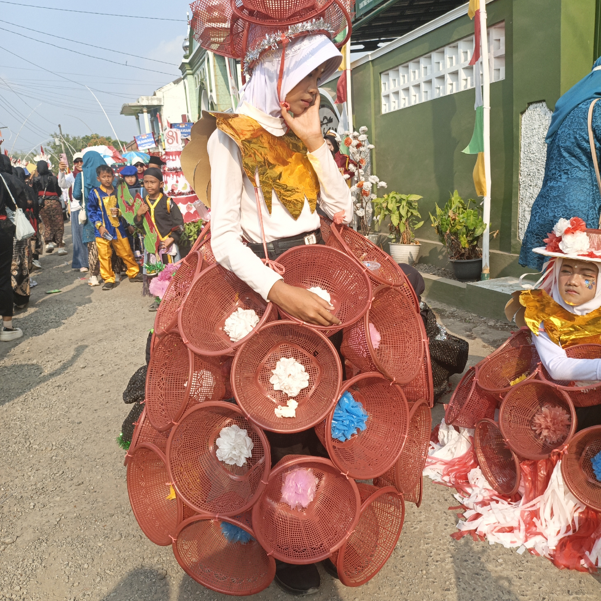 In the city of Tanahbaya, Indonesia, on a sunny day, a festive parade is underway. The central figure is a young person adorned in an elaborate costume. They wear a white hijab and a gold, feather-like collar. Their body is encased in a spherical structure made of interconnected red plastic baskets, some containing white and blue decorative flowers. A large, red, basket-like headdress sits atop their head, adorned with silver tinsel. The individual is captured mid-gesture, with their hand touching their chin, appearing thoughtful.
To their right, another person, also in costume, is partially visible. This individual wears a white hijab, a gold collar, and a hat decorated with red and white flowers. Their face is also painted with blue cosmetic embellishments around the eyes.
In the background, a procession of people is walking along a dirt road. Children in colorful traditional attire, including bright blue and yellow garments, are visible. Some individuals are carrying flags and banners. The buildings flanking the road are painted green, with white decorative grilles. Green plants in pots are placed near the buildings. The ground is covered in gravel and dirt. The overall atmosphere suggests a lively cultural event or celebration.