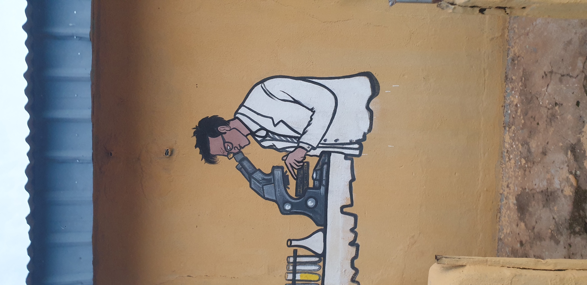A mural depicts a person wearing glasses and a lab coat, hunched over a microscope. To the left of the microscope are several test tubes containing yellow liquid and a conical flask. The mural is painted on a yellow wall beneath a corrugated metal roof. The image is taken at an angle from below, looking upwards. The location is Sabon Tasha, Nigeria.