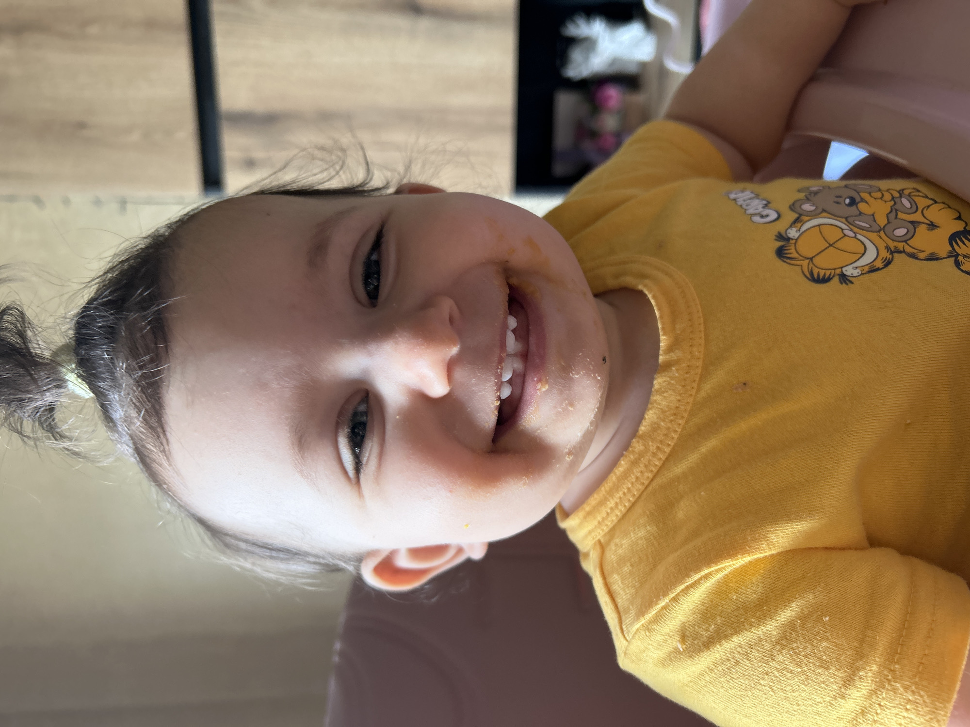 The image displays a close-up, angled portrait of a cheerful baby, likely between 6 to 18 months old, captured indoors. The baby is smiling broadly, revealing a few front teeth, and looking directly at the camera. Their face and the area around their mouth are smudged with yellowish food particles and crumbs, indicating they have recently eaten or are in the process of eating.
The baby has dark hair, styled in a small bun or ponytail on one side. They are wearing a vibrant yellow t-shirt that features a cartoon print. The print depicts two characters: one clearly recognizable as Garfield the cat, and another, possibly a bear or dog, resting on Garfield's back. The word "GARFIELD" is partially visible as text on the shirt.
The background suggests an indoor home setting, with a light wooden floor or wall visible. In the upper right corner, blurry elements include what appears to be a white tissue box and a pink or light-colored object, possibly part of a high chair. The bright, even lighting indicates the photo was taken during the daytime. The baby's expression radiates joy and playfulness, conveying a happy moment. The image is oriented with the baby's head to the right, suggesting a 90-degree clockwise rotation from a typical upright view.