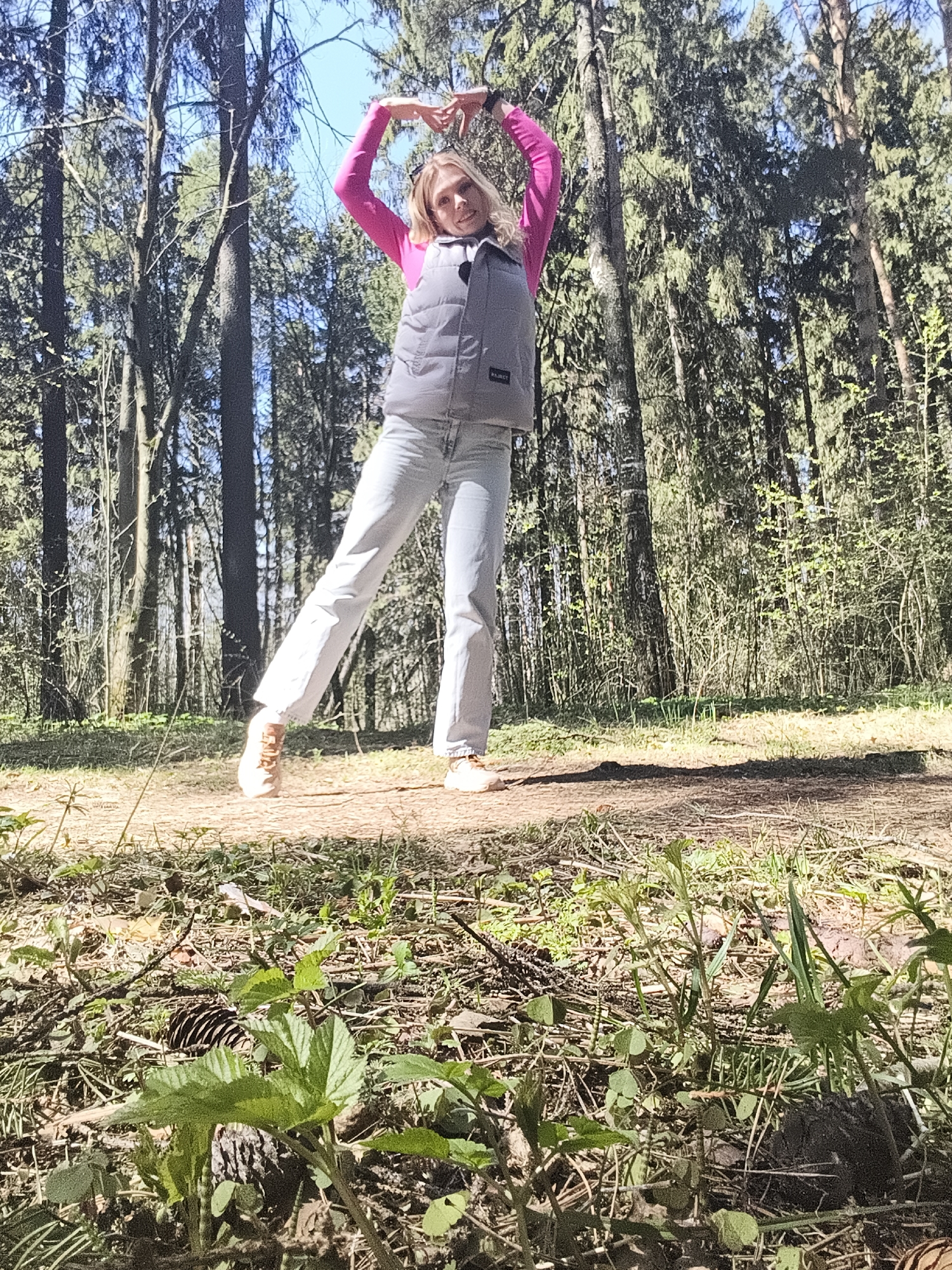 A fair-skinned woman stands in a forest, likely in Izhevsk, Russia. She wears a bright pink long-sleeved shirt, a gray vest, light blue jeans, and tan shoes. Her arms are raised above her head, forming a heart shape with her hands. The forest background consists of tall trees, some with visible sunlight shining through. The foreground is covered in forest debris including leaves, pine needles, small sprouts, and twigs.