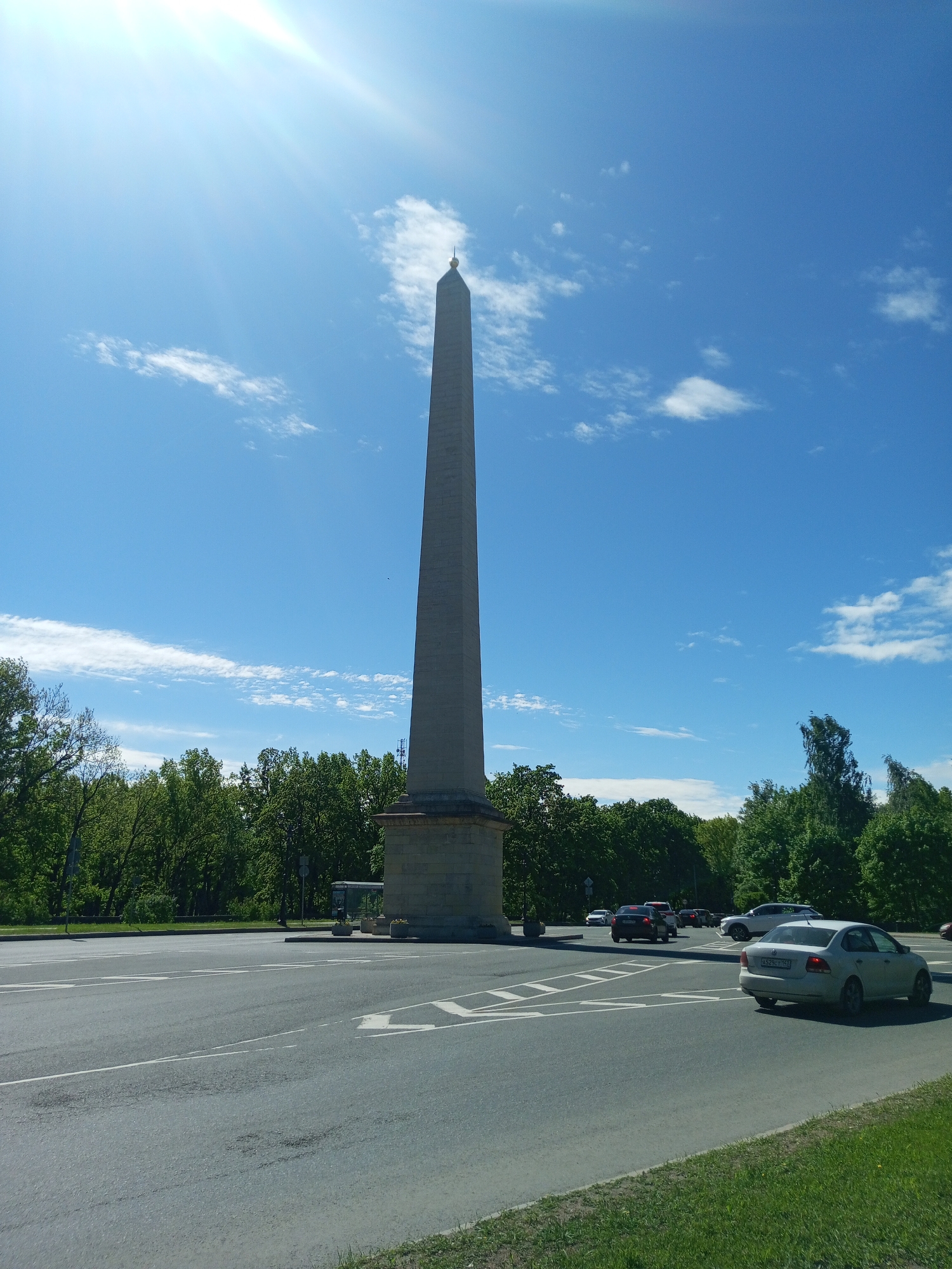 The image shows the Obelisk of Glory in Gatchina, Russia, a tall, gray stone obelisk on a raised platform. The obelisk is located at a road intersection with several cars visible, including a white sedan in the foreground. Trees line the background, and the sky is bright blue with scattered white clouds. The sun is shining brightly, causing glare in the upper left corner. A bus stop is located near the obelisk.