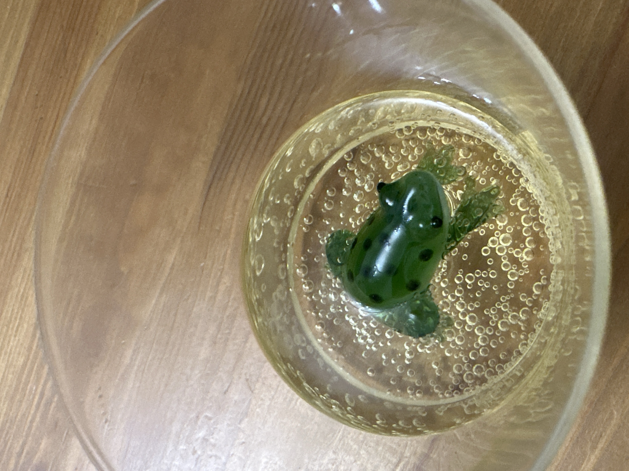 A glass of yellow liquid containing a green, frog-shaped candy is displayed on a wooden surface. The liquid is effervescent, with numerous small bubbles dispersed throughout. The candy, which resembles a frog, is partially submerged in the liquid. The wooden surface appears to be a tabletop with a natural wood grain. The image is taken from a top-down perspective, focusing on the glass and its contents. There are no people or other objects visible in the scene. The lighting suggests an indoor setting, likely natural light from a window. The overall impression is one of a still life, with a playful and whimsical element introduced by the candy.