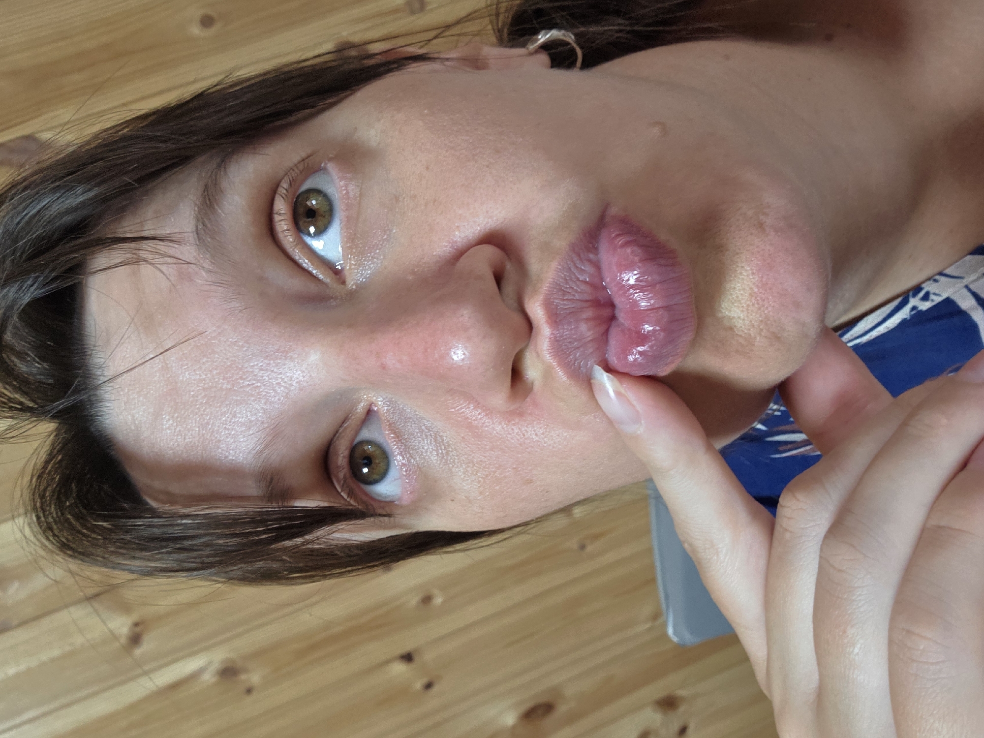 The image shows a close-up of a woman's face, with her mouth pursed and a finger touching her lips. She has brown hair and brown eyes, and is wearing a silver hoop earring. The background is a wooden wall. There is no visible indication of the city of Dunnae-myeon, South Korea. The image is a selfie or a self-taken photograph.