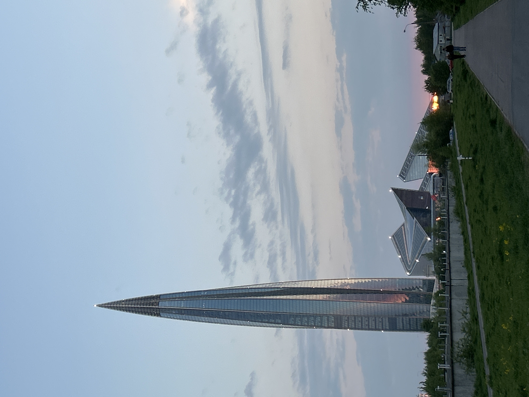 The image shows the Lakhta Center skyscraper in Saint Petersburg, Russia, prominently featuring its spire-like structure against a backdrop of a light blue sky with wispy clouds. The building's glass facade reflects the sky and sunset colors. The base of the skyscraper leads to smaller structures along a concrete walkway bordered by green vegetation. Two individuals are walking on the walkway, and there are visible lampposts and street elements, suggesting a public or commercial area. The scene conveys a blend of urban architecture and natural elements under an evening light.
