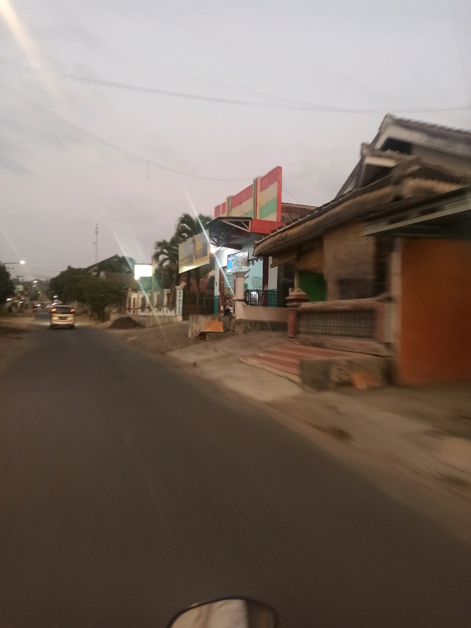 The image depicts a street scene in Tlogosari, Indonesia, at dusk. A paved road occupies the foreground, curving slightly to the left. On the right side of the road, buildings with varied architectural styles are visible. One building features a textured brown facade and an orange-colored side. Another structure has a multi-colored facade with red, green, and white panels. A white railing and a small bench are also present near the buildings.
To the left of the road, a light-colored car is driving away from the viewer. In the distance, more buildings and trees line the street, and streetlights are illuminated. The sky is overcast, with a pale glow suggesting the fading light of day. A telephone pole is visible further down the road. The overall impression is that of a residential or mixed-use area experiencing the transition from day to night.